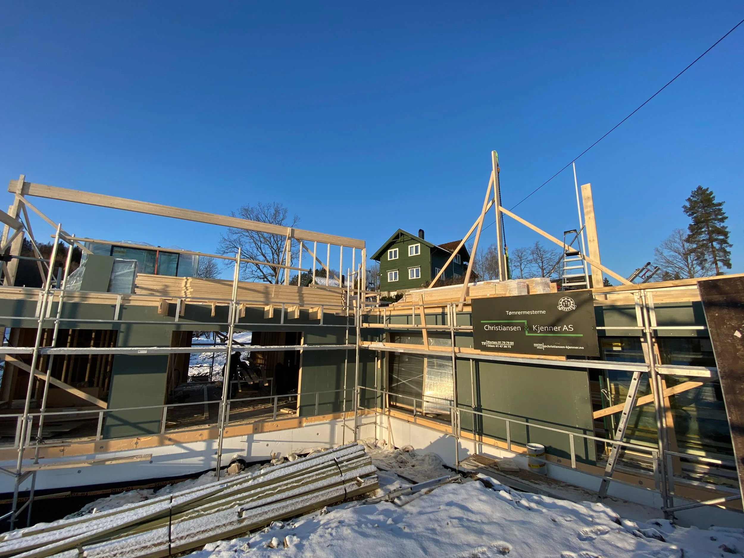 Påbegynt bygge med treverk og stillaser i snø, under en klar blå himmel, med hus i bakgrunnen.