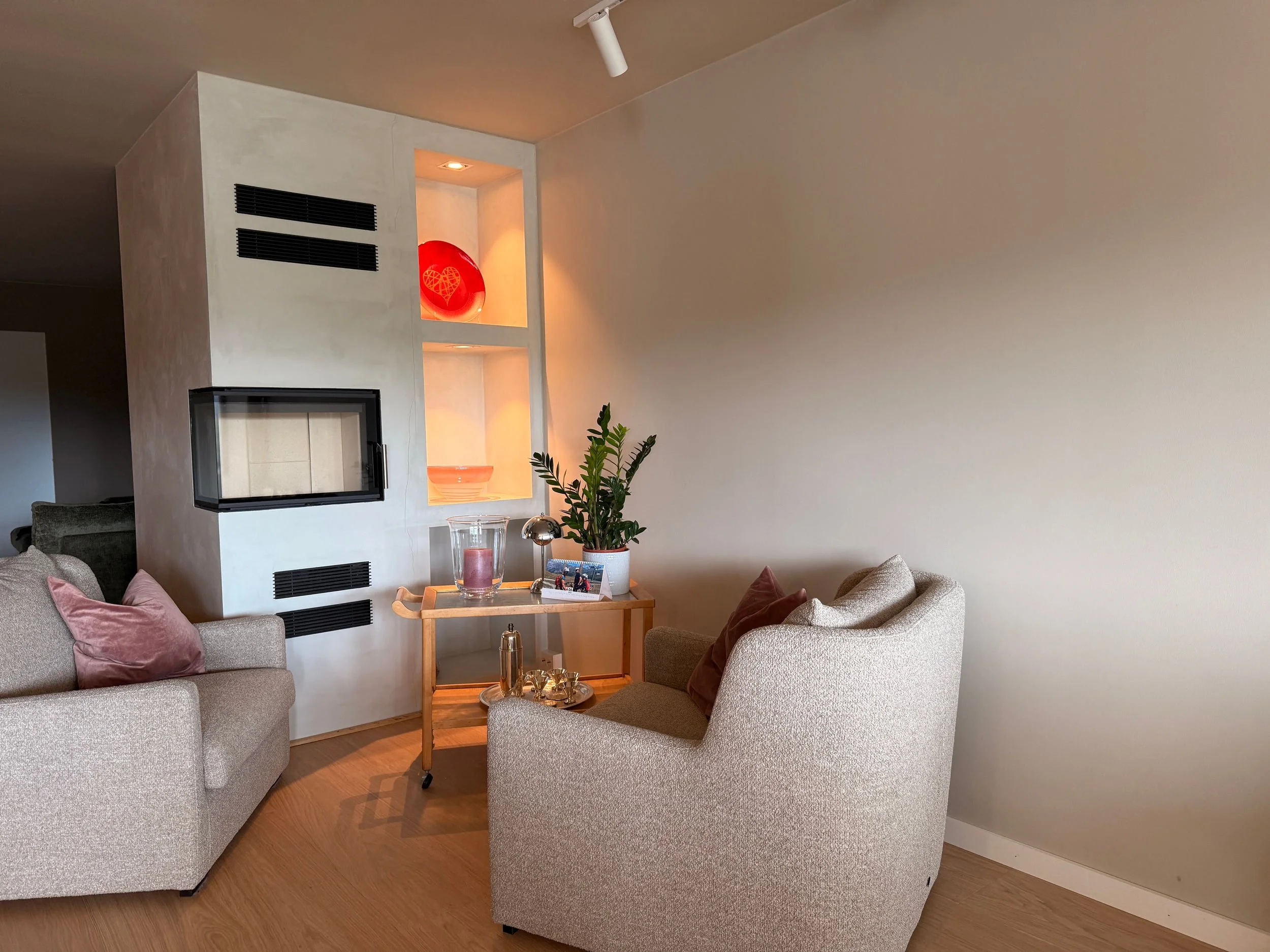 Et koselig stuehjørne med to beige lenestoler med puter, en trebjelle med pynt, en liten plante, og en glassvase med lys ved siden av et vegghengt elektrisk peis, med en trappestige med to hyller som inneholder dekorative ting.