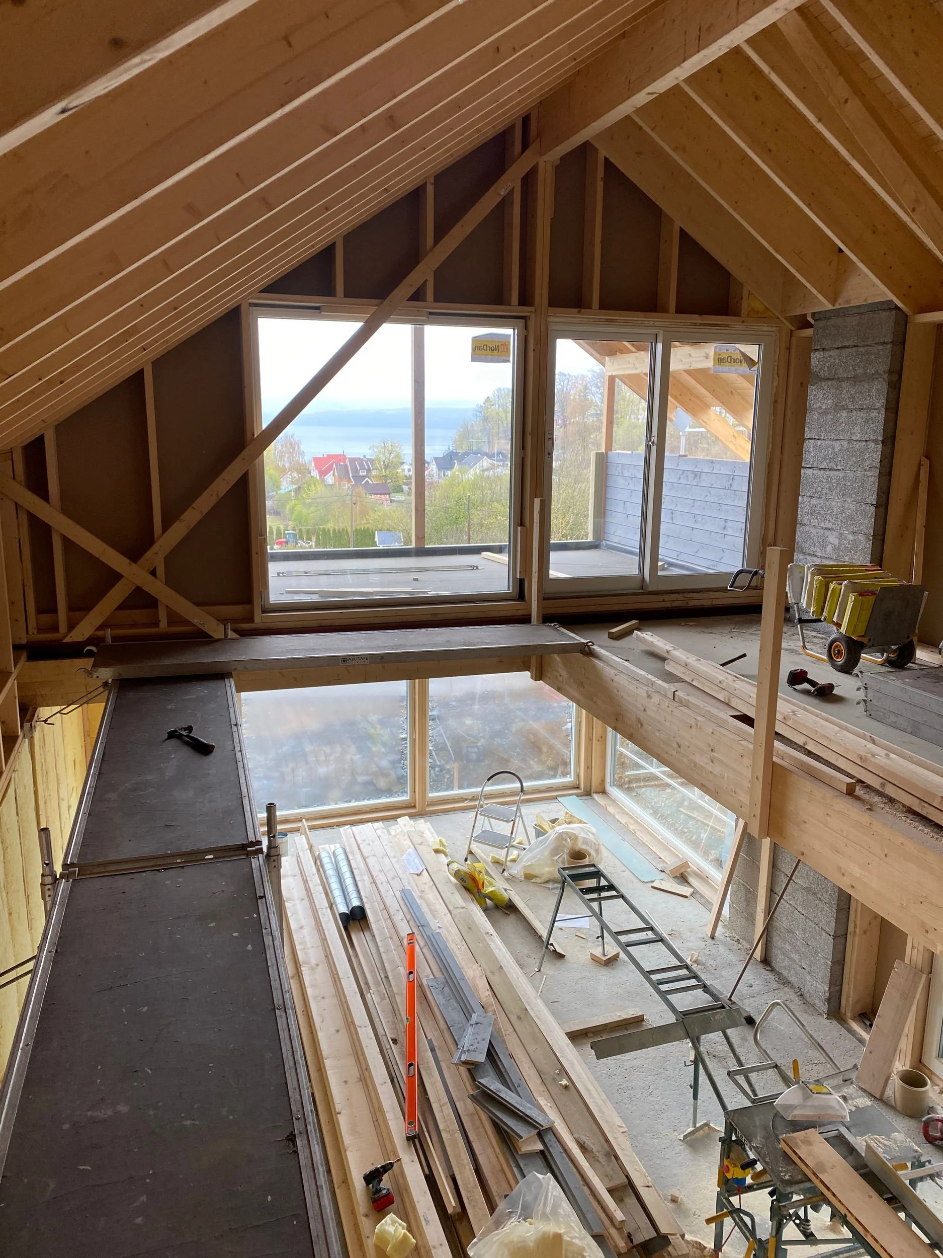 Innendørs bygging med store vinduer, treverk og byggeutstyr. Uthus under konstruksjon med scaffoldings og materialer på gulvet.