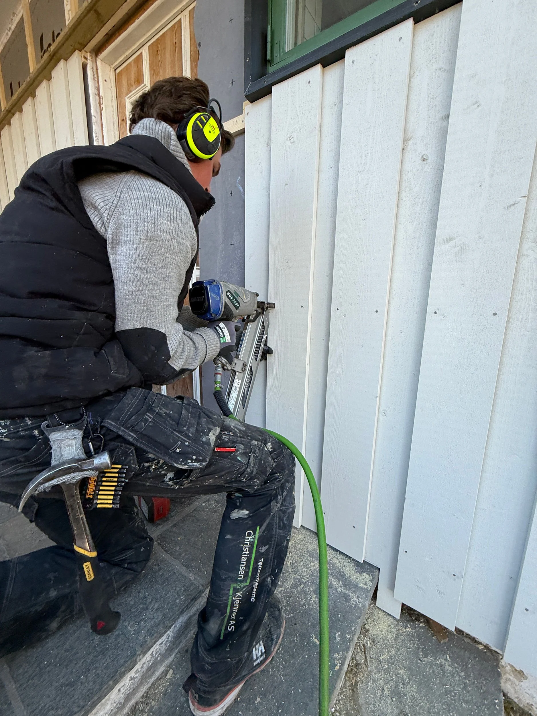 En person bruker en elektrisk verktøy til å montere eller bore inn i en hvit vegg utendørs. Personen har på seg hørselsvern, arbeidsklær og verktøybelte. Det er snø på bakken rundt.