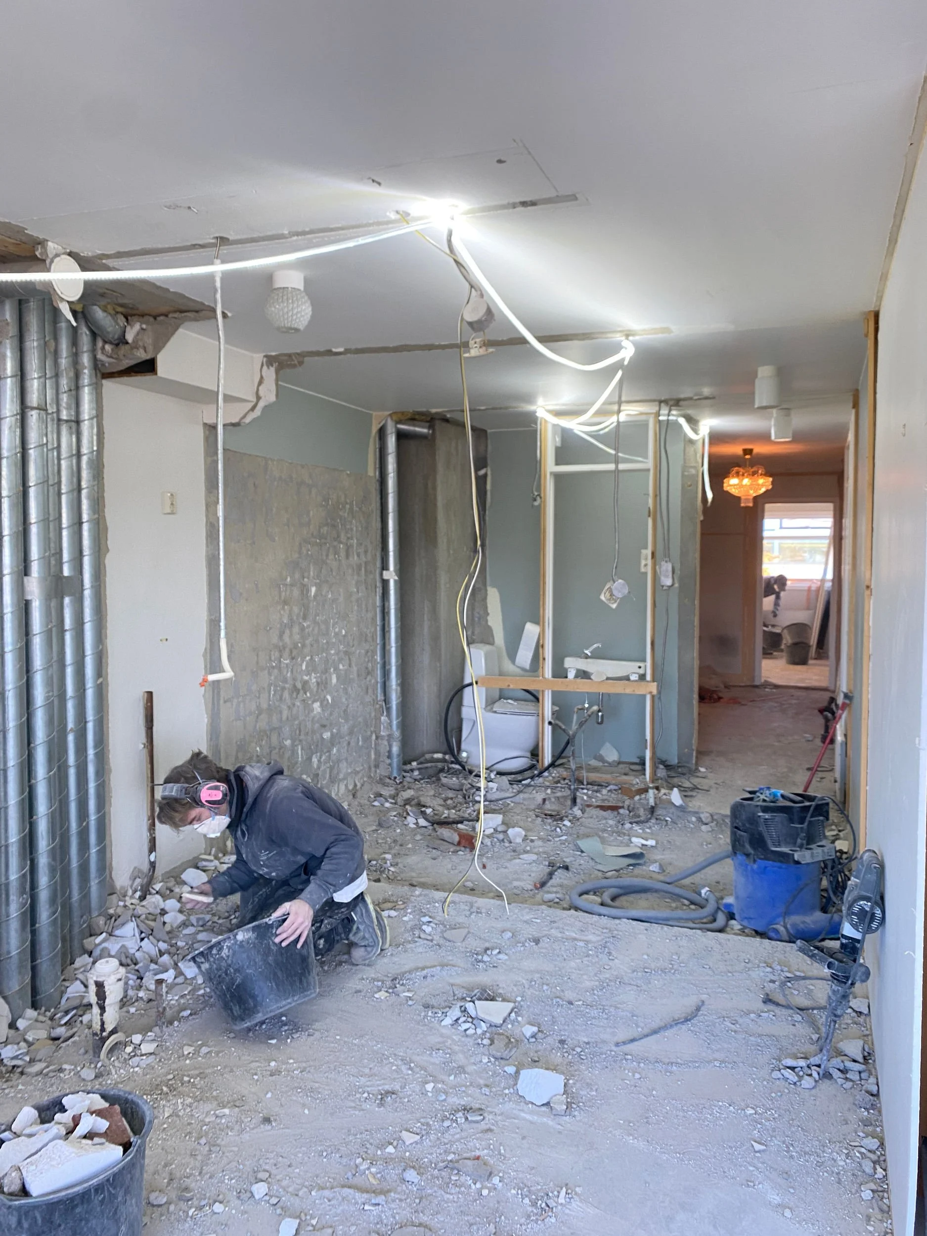 En person som jobber med renovering innendørs, omgitt av byggeavfall, rør og verktøy.