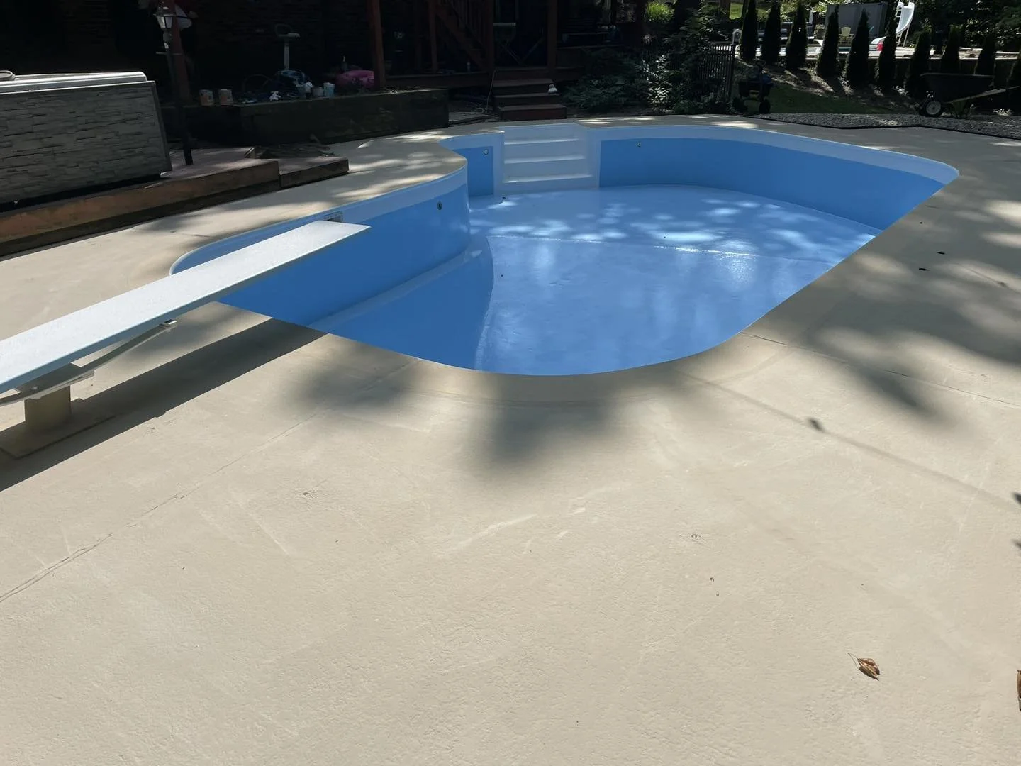 Empty backyard pool with a blue interior and attached slide, surrounded by a concrete deck, with outdoor furniture and trees in the background.