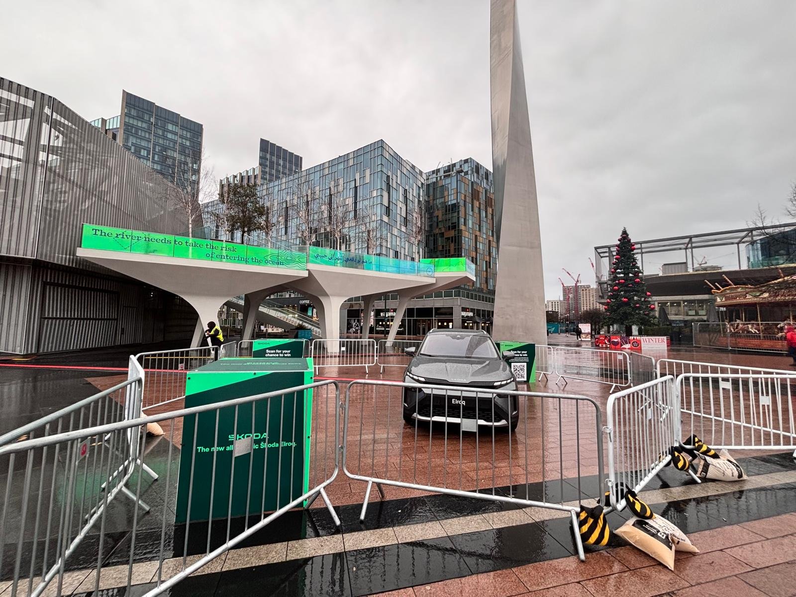 An outdoor urban scene on a rainy day includes a Christmas tree decorated with red ornaments, modern glass buildings, a small electric car, and temporary barriers of heras fecing around the area. There's also large digital display showing green text