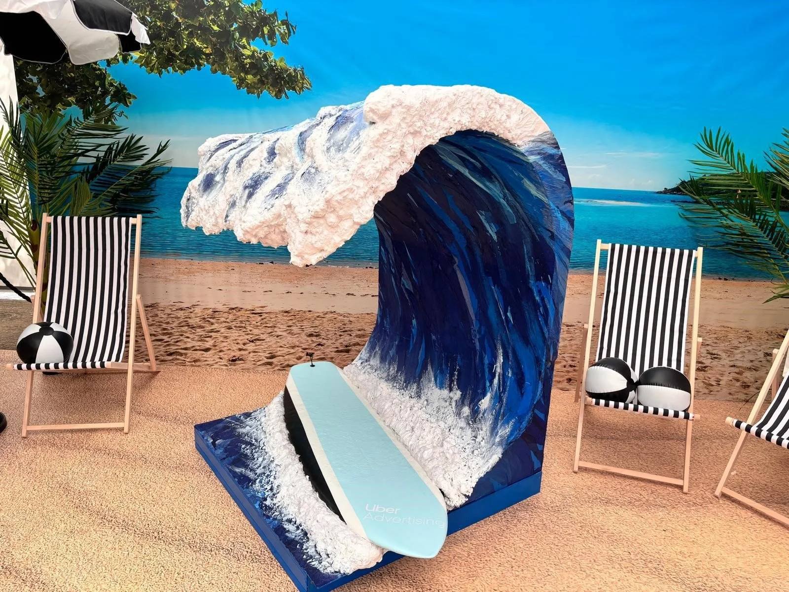 Indoor beach-themed display with a large wave sculpture, beach chairs with striped fabric, a surfboard, and tropical plants, set against a backdrop of a beach and ocean.