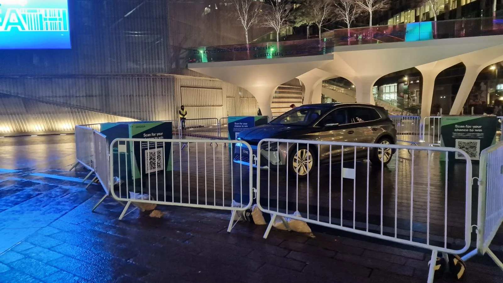 A black electric car parked behind heras fencing at night, with QR code scanning stations nearby, against a modern architectural building with illuminated trees in the background.