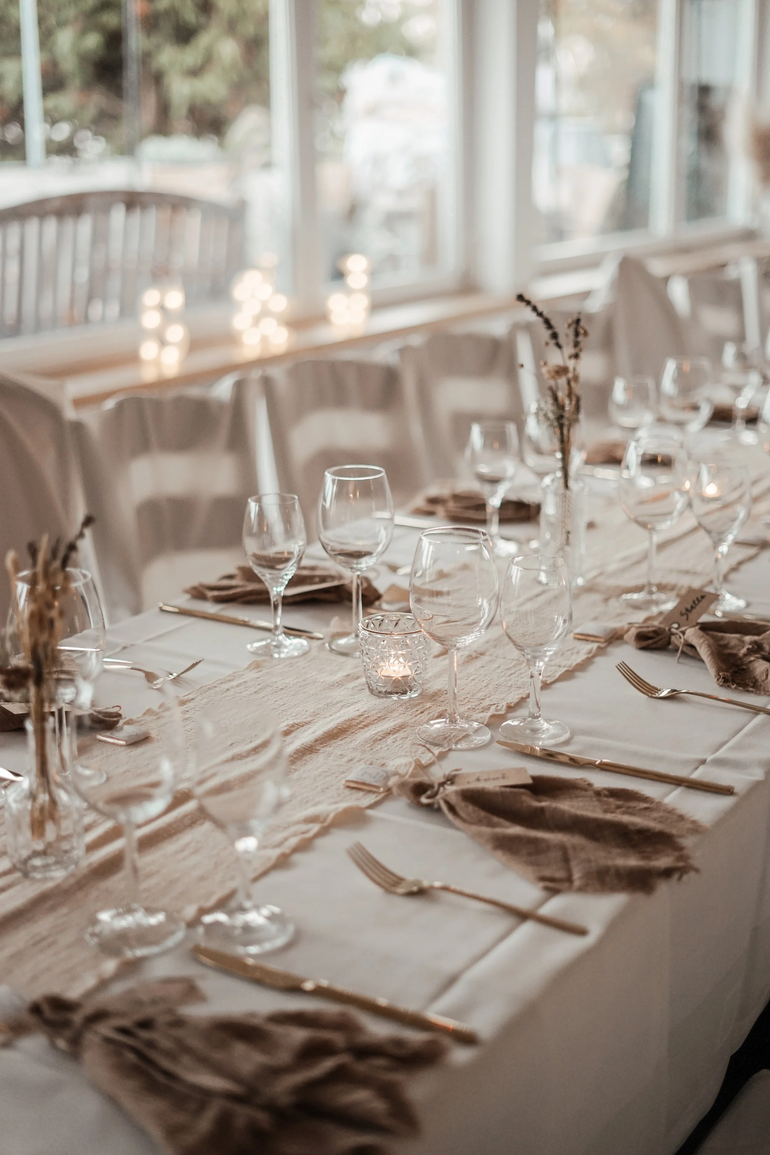 Ein festlich gedeckter Esstisch mit Wein- und Wassergläsern, Kerzen in Gläsern und einer Mitteldecke in neutralen Tönen, vor einem Fenster mit Blick im Hintergrund.