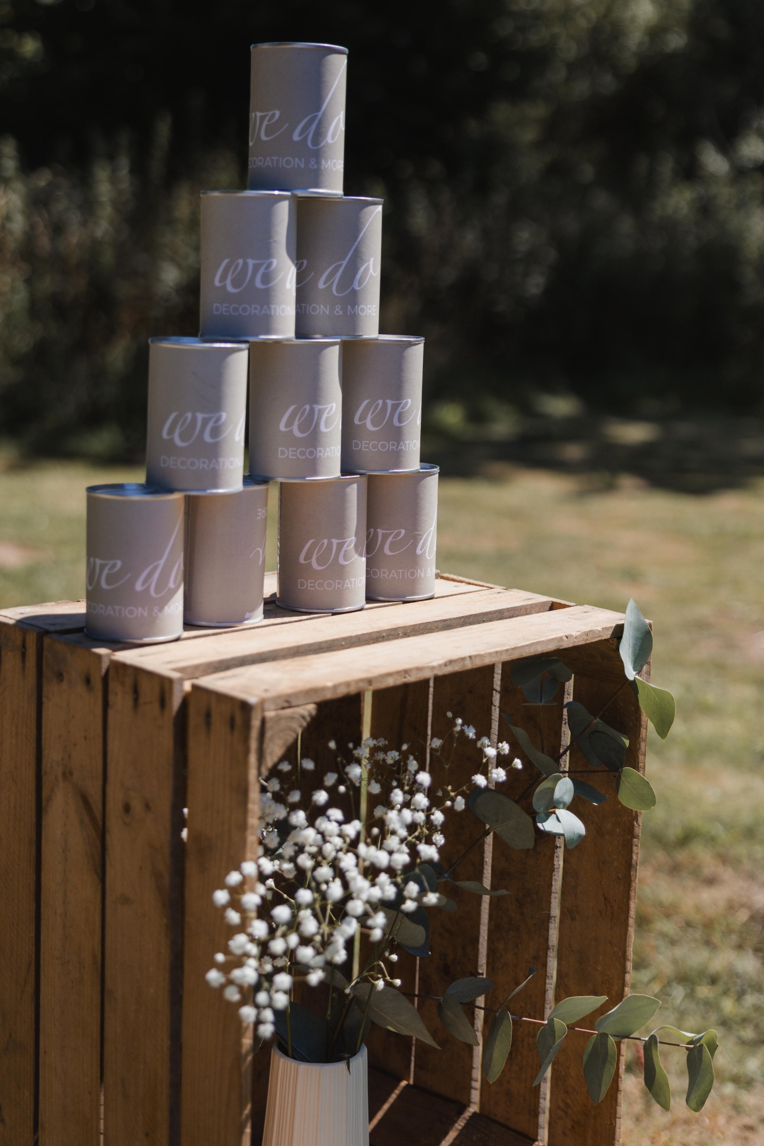 Weiße dekorative Dosen mit Schriftzug 'we do' gestapelt auf einer Holzkiste, darunter eine Vase mit weißen Blumen und grünen Blättern im Freien.