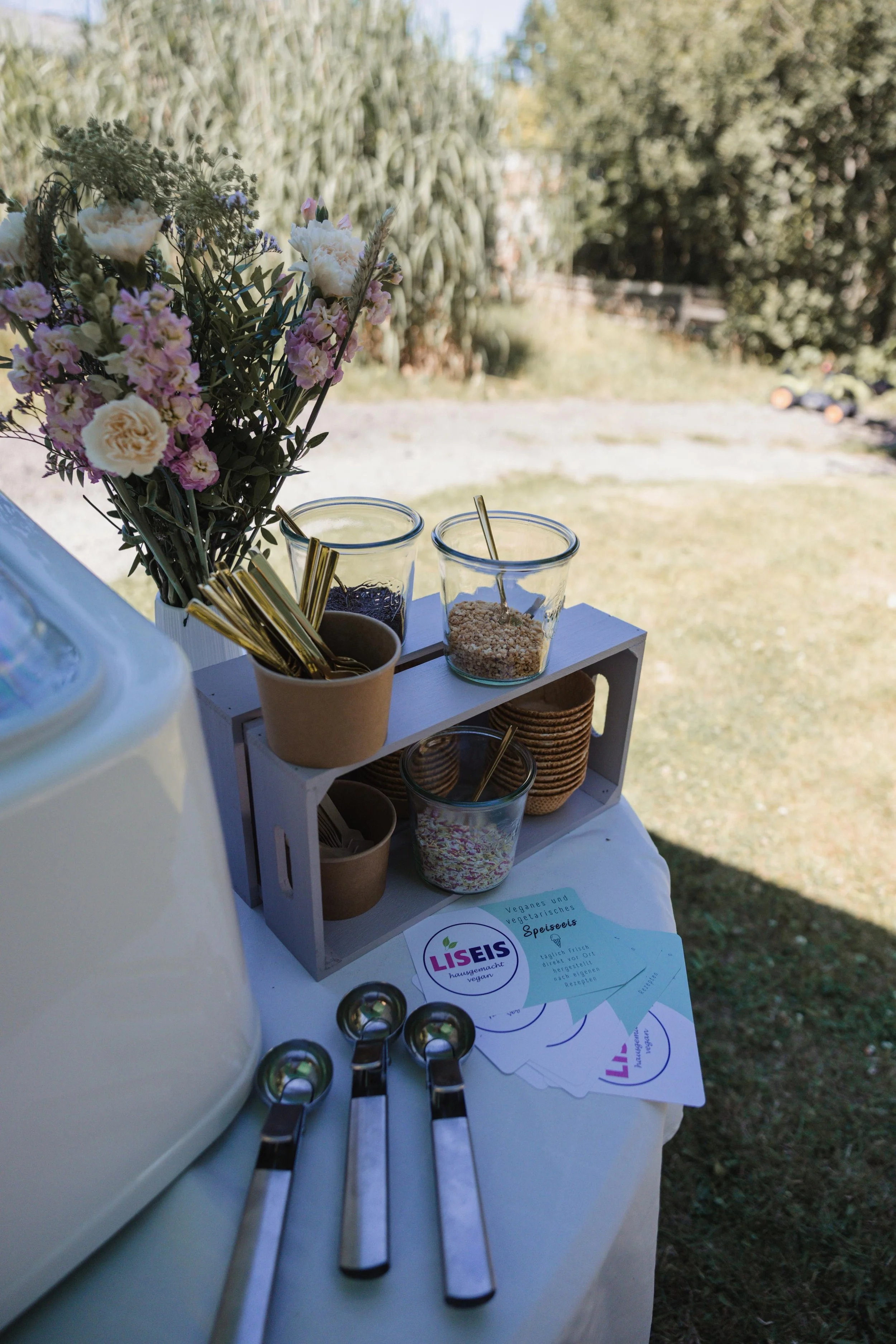 Essensstation mit verschiedene vegane und vegetarische Zutaten in Gläsern, ein Blumenstrauß und Löffel auf einem weißen Tisch im Freien.