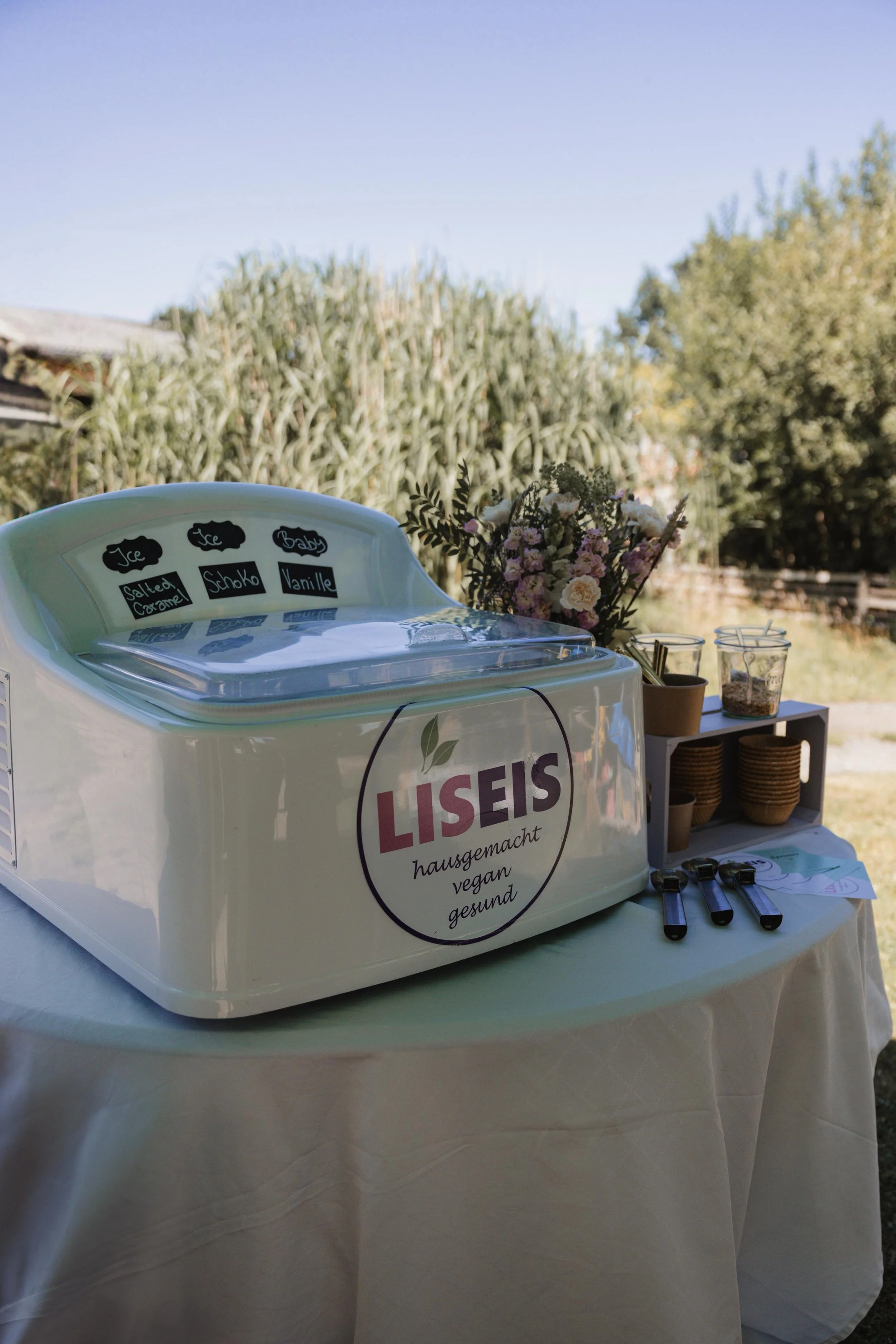 Ein weißer Eiswürfelspender mit einem Logo von Liseis, im Hintergrund ein Blumenstrauß und eine Tierschale, auf einem weißen Tischtuch im Freien, umgeben von Bäumen und Himmel.
