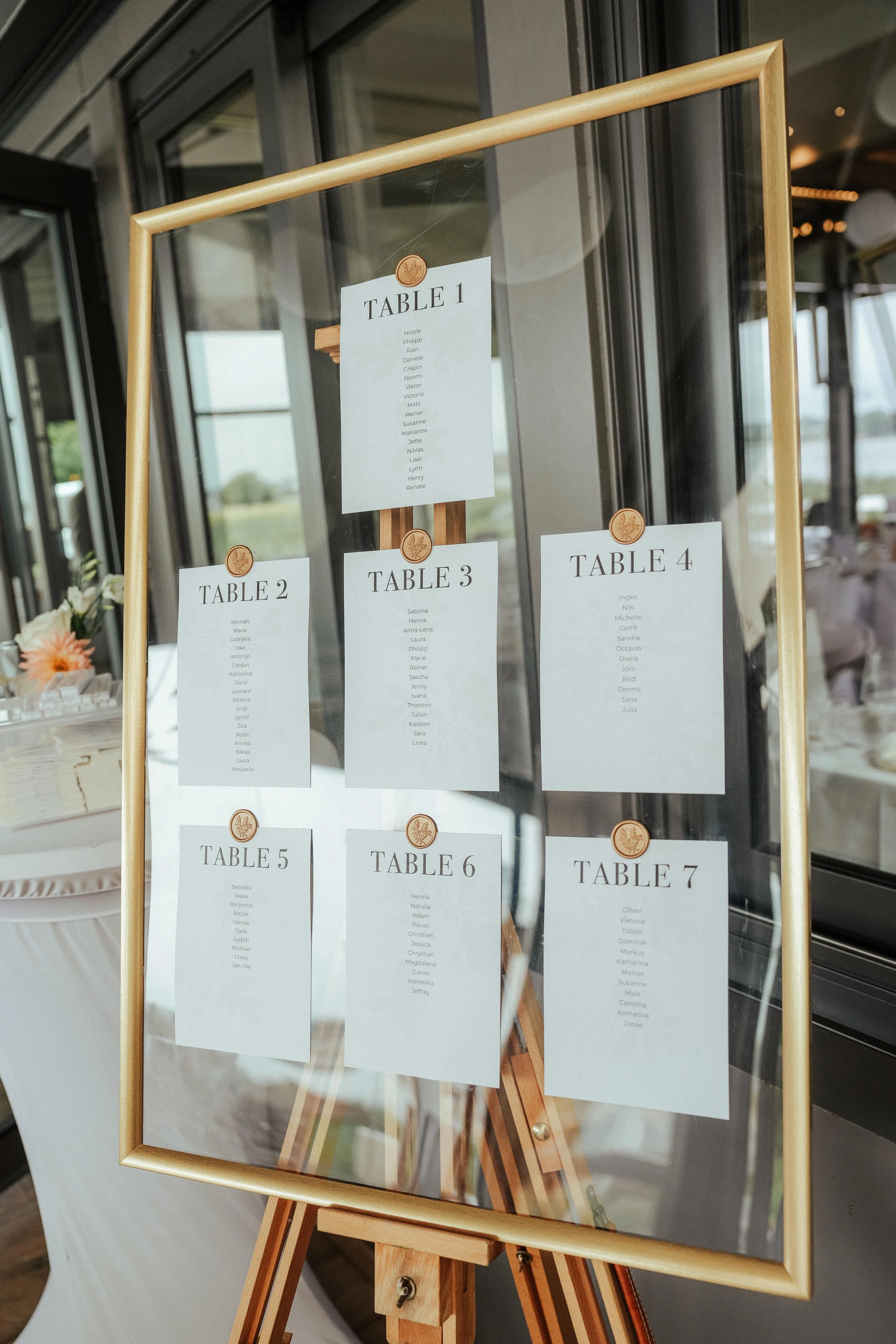 Sitzplan für eine Veranstaltung mit sieben Tischen, jeder mit einer Liste von Namen, auf einem Holzrahmen, reflektierendem Glas, mit einem modernen Raum im Hintergrund.