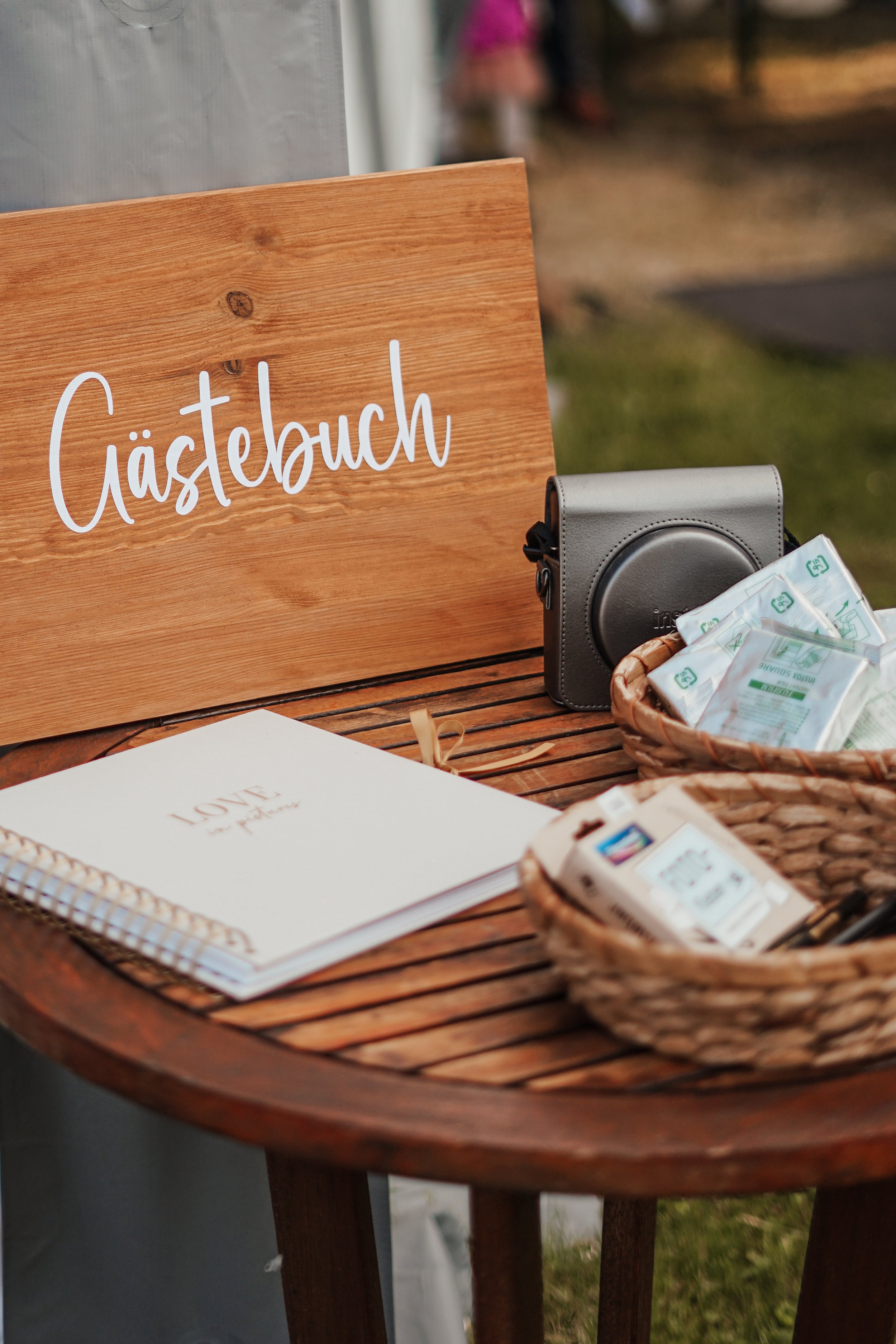 Eine Holztisch mit einem Schild mit der Aufschrift 'Gästebuch', einem Notizbuch, Kameratasche, Körbe mit Papier und Stiften, vermutlich bei einer Gästebuch-Station im Freien.