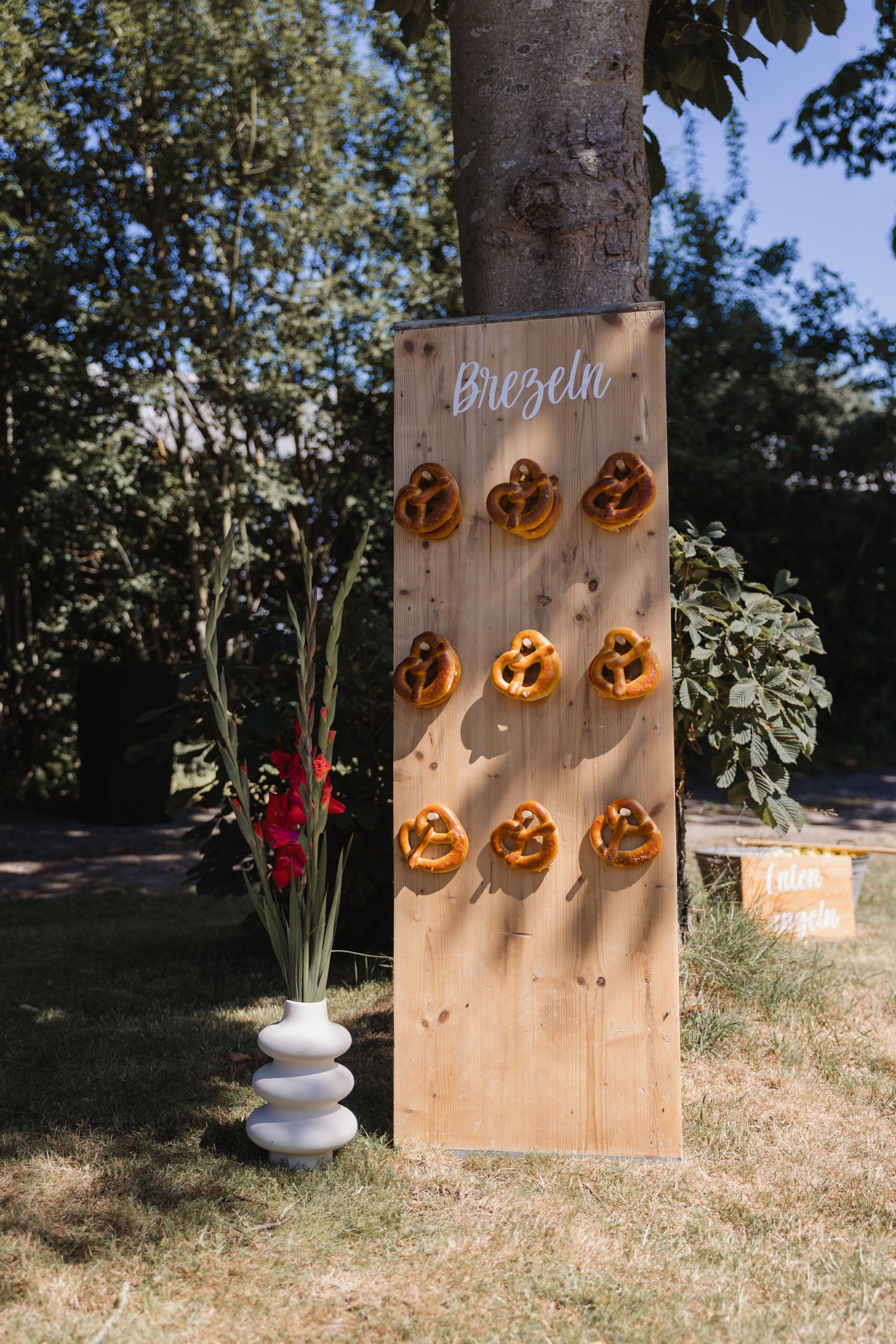 Holztafel mit dem Schriftzug "Brezeln" und mehreren Brezeln, die an der Tafel befestigt sind, im Freien mit Bäumen und Himmel im Hintergrund.