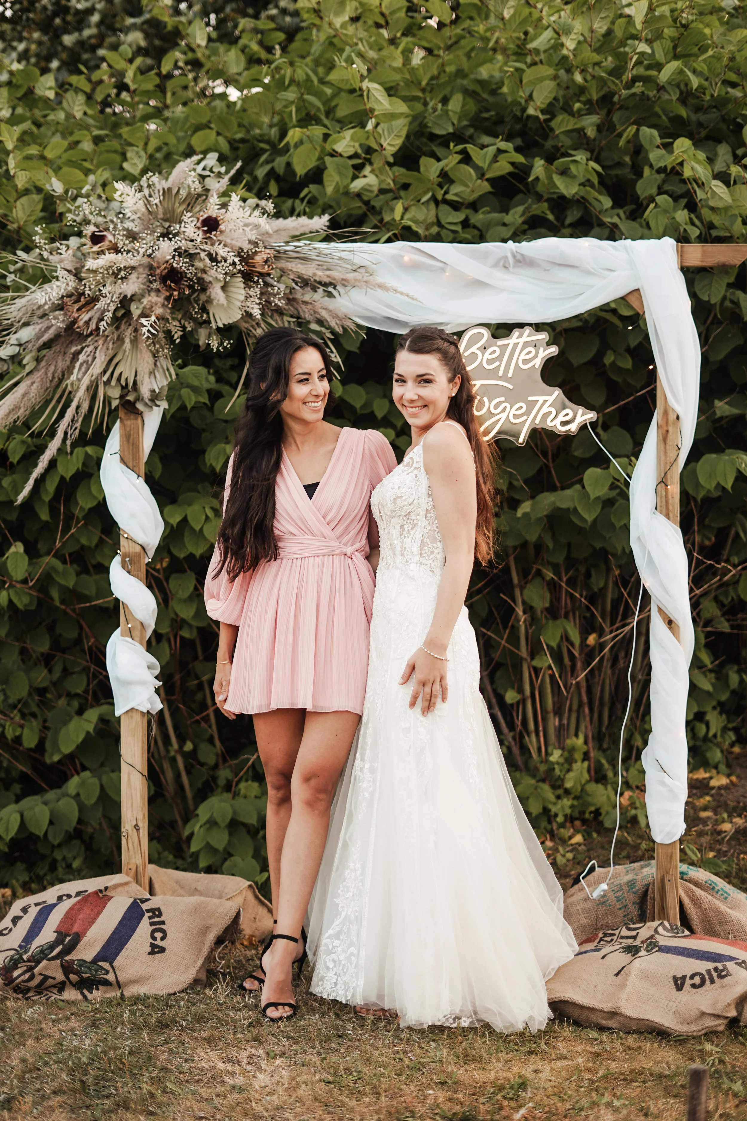 Zwei Frauen stehen unter einem dekorativen Bogen mit einem Blumenarrangement und einem Neon-Schild mit der Aufschrift "Better Together" bei einer Hochzeit im Freien.