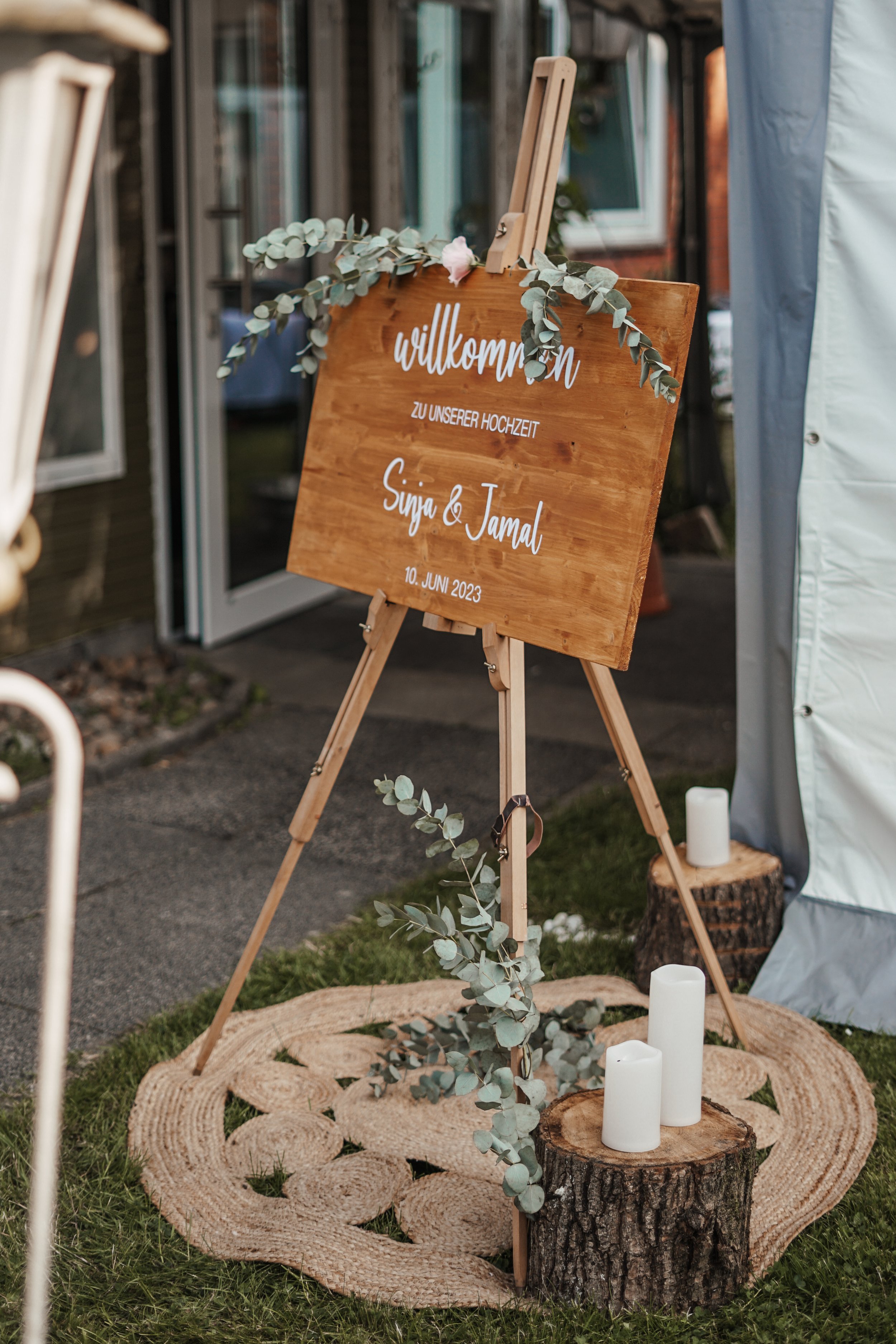 Ein hölzernes Willkommensschild mit weißen Schriftzügen für eine Hochzeit, dekoriert mit Eukalyptus, steht auf einem Holzständer, umgeben von Kerzen auf Baumstümpfen, vor einem Zelt. Es trägt die Aufschrift: "willkommen zu unserer hochzeit, Sina & Jamal, 10. Juni 2023."