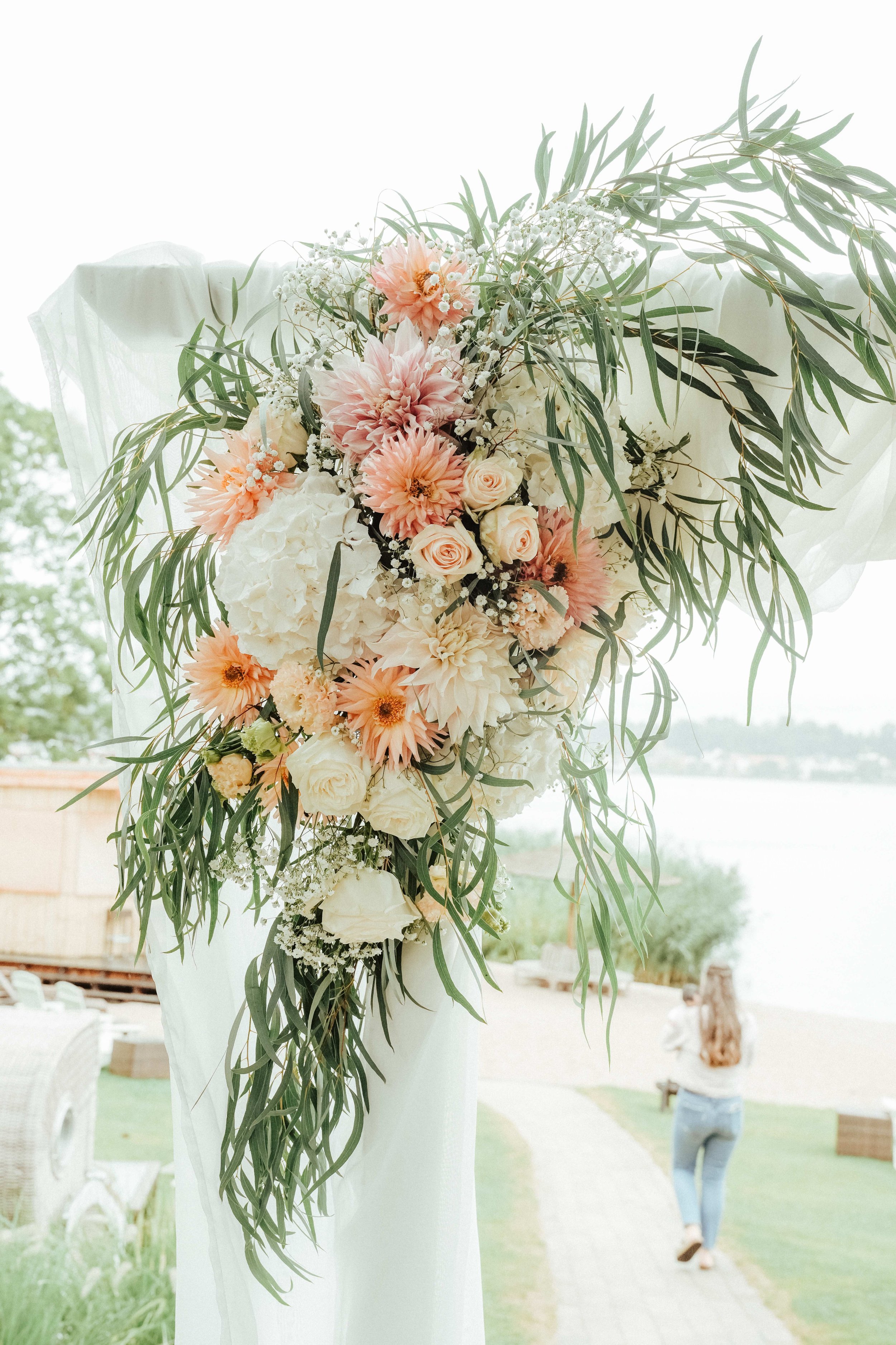 Blumenbogen mit weißen und peachfarbenen Blumen, darunter Rosen und Dahlien, vor einem Wasser im Hintergrund bei einer outdoor Veranstaltung.