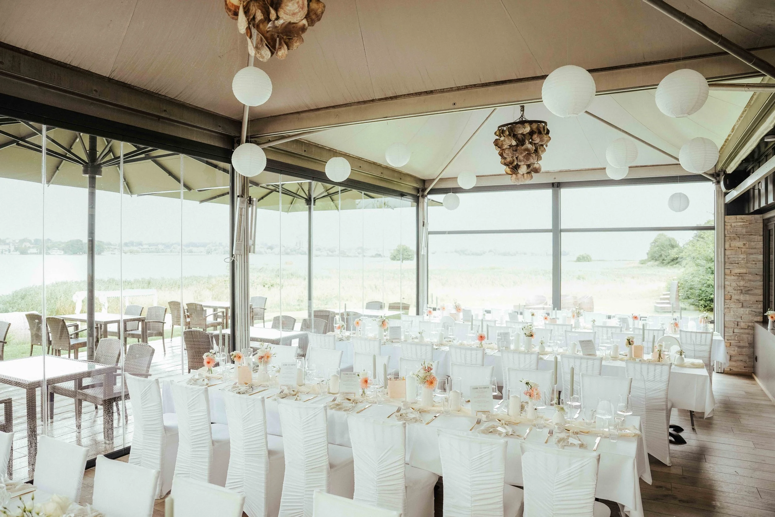 Ein festlich dekorierter Veranstaltungsraum mit weißen Tischen, Stühlen, Blumenarrangements, Papierlampen und Blick auf eine Wasserlandschaft im Hintergrund in Kiel und Umgebung