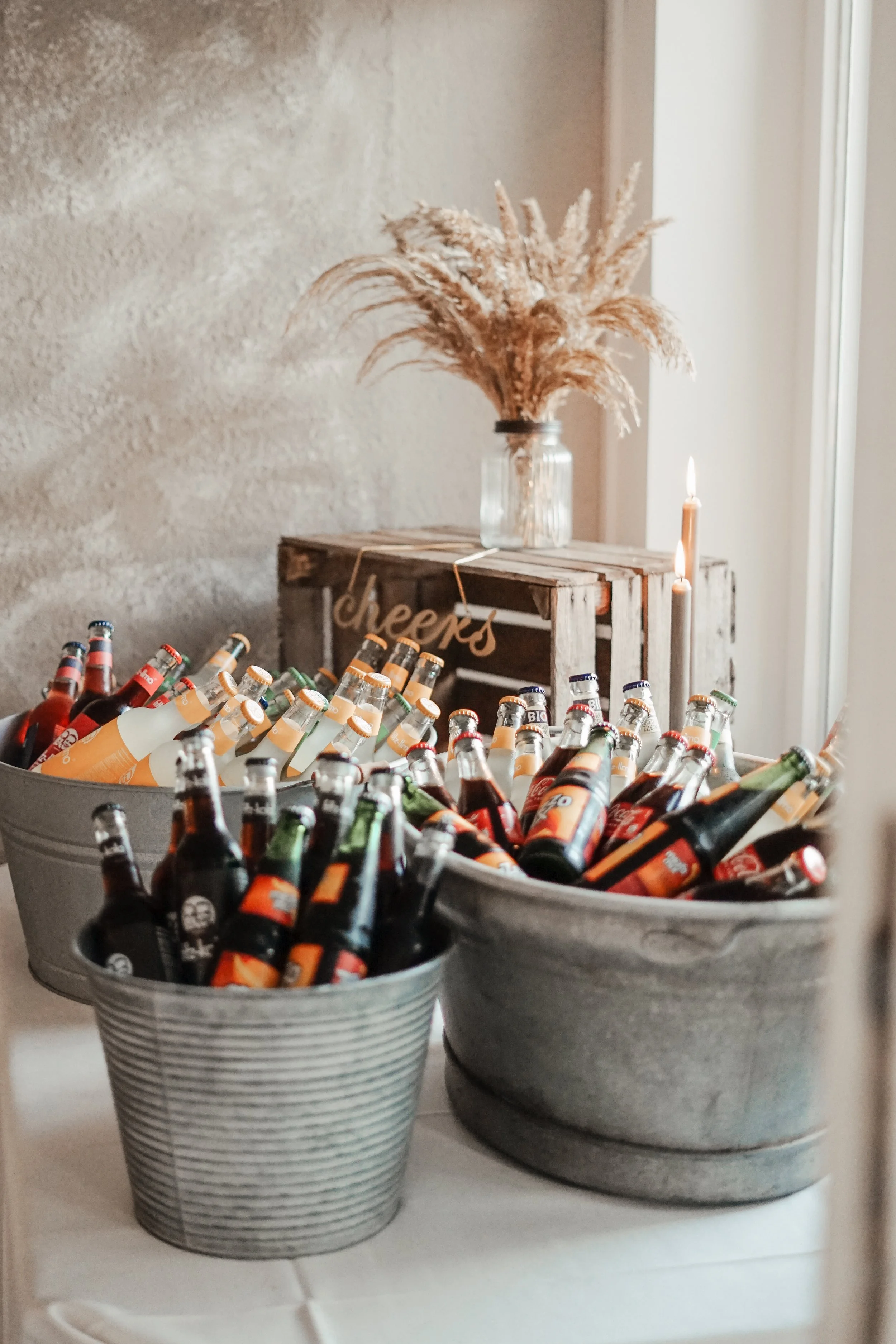 Eisengläser mit verschiedenen Bierflaschen in zwei großen Körben, Deko im Hintergrund mit Topf und getrockneten Pflanzen, Kerzen, Holzbox, Beschriftung 'Cheers'.
