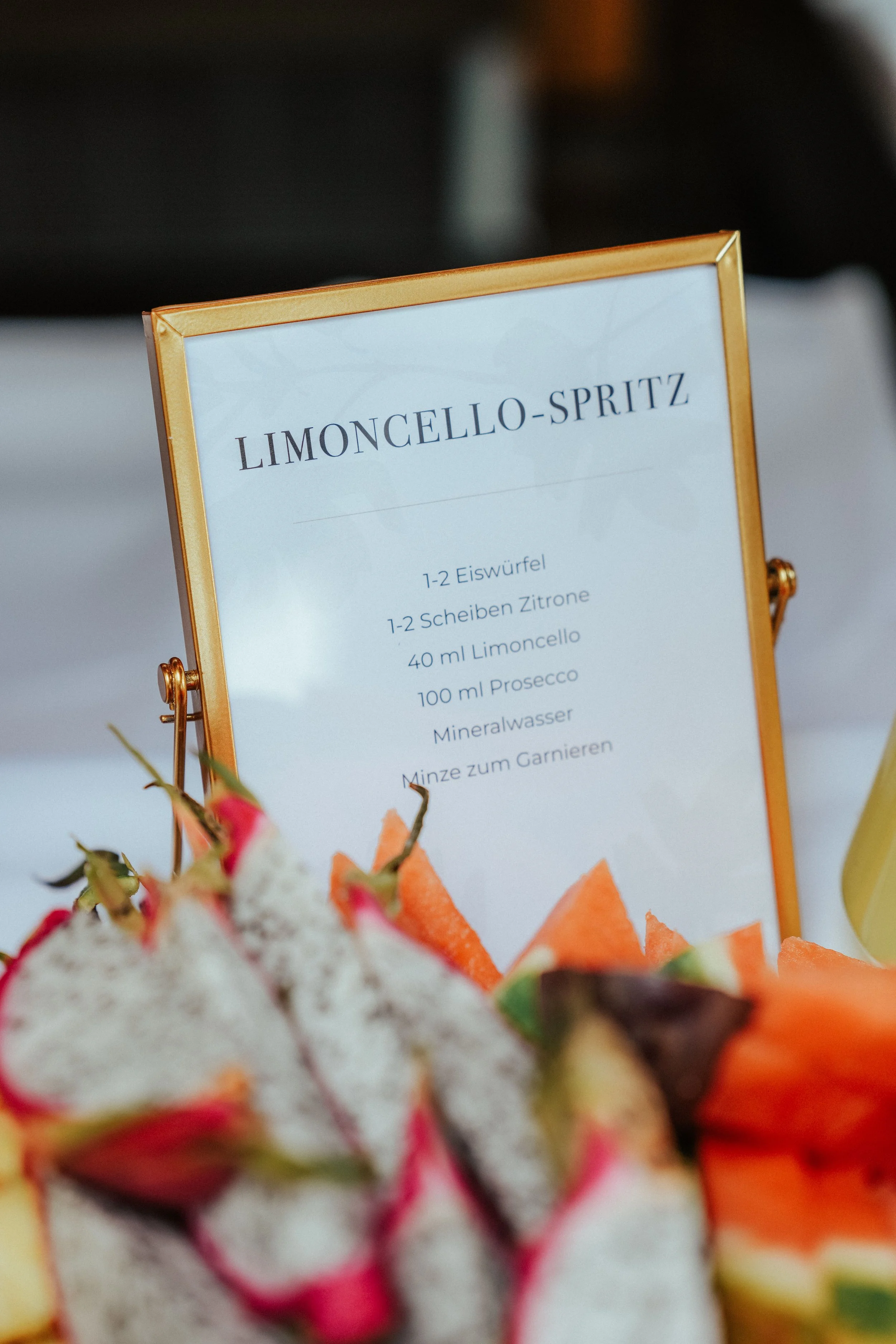 Ein goldgerahmtes Menü mit dem Titel 'LIMONCELLO-SPRITZ' und den Zutaten, darunter Eiswürfel, Zitronenscheiben, Limoncello, Prosecco, Mineralwasser und Minze. Im Vordergrund sind frische Früchte, darunter Erdbeeren und Melone, sichtbar.