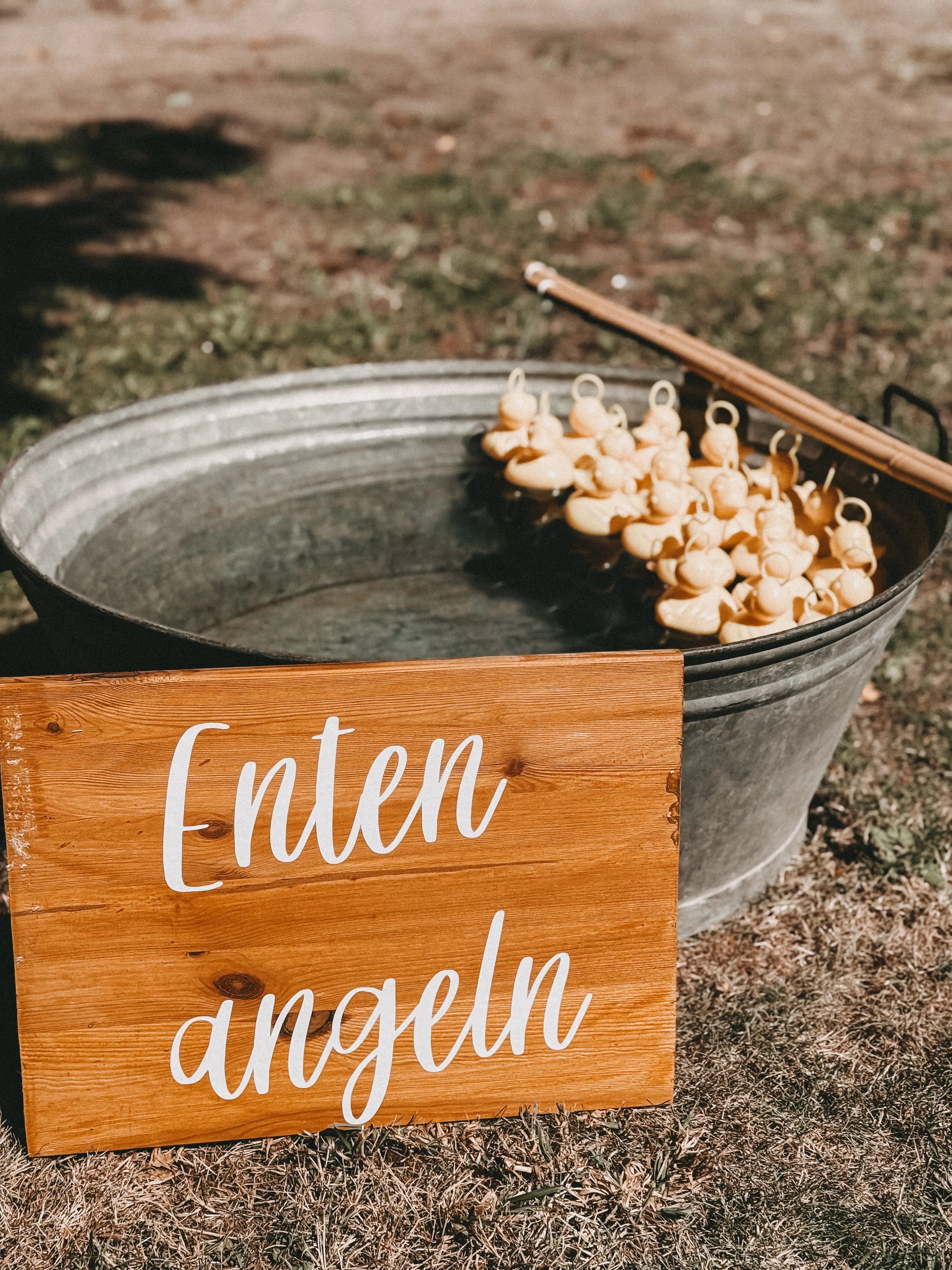 Ein Eimer mit Holzherzen und einem Schild mit der Aufschrift 'Enten angeln' auf dem Boden im Freien.