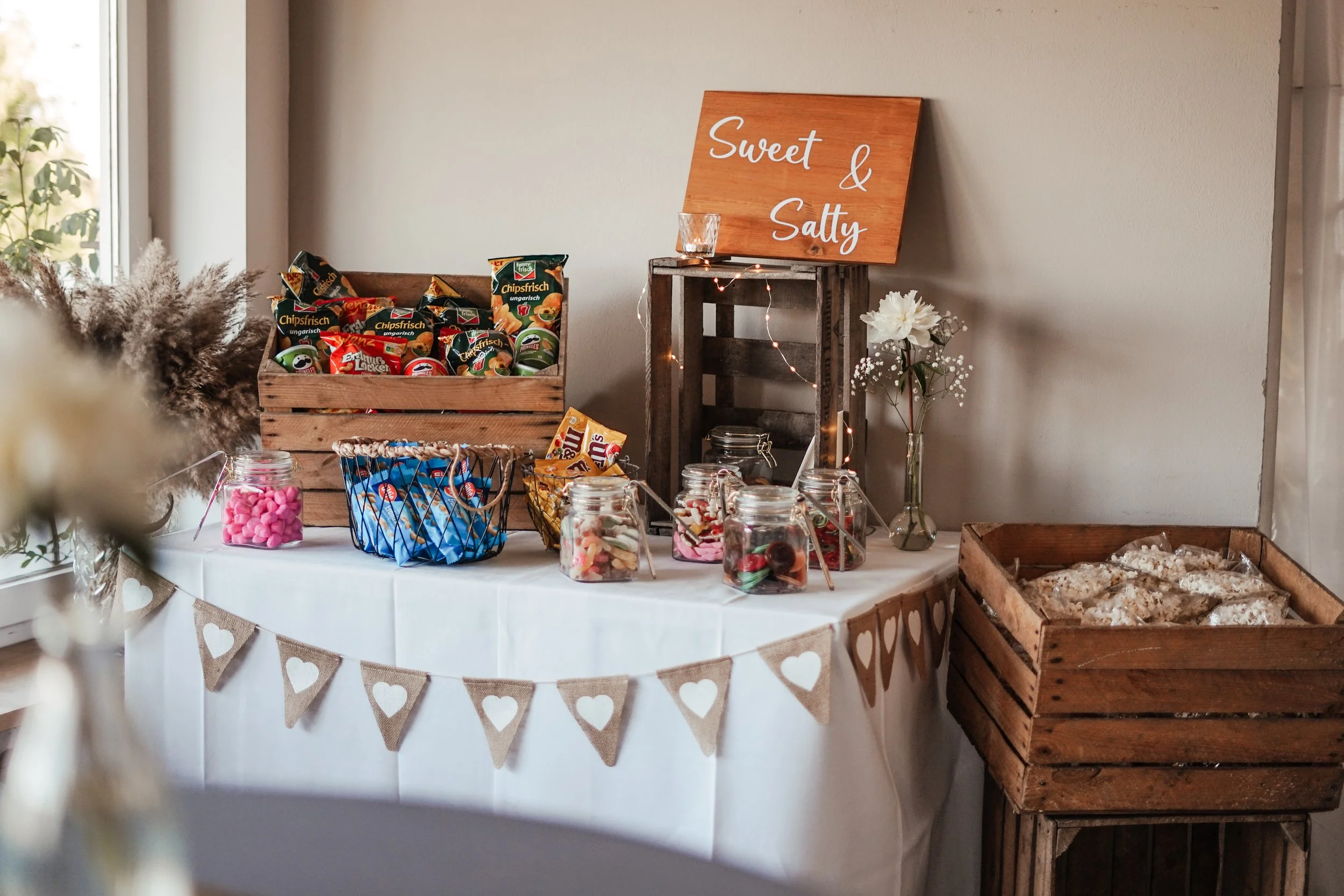 Tisch mit Süßigkeiten und Snacks, Dekoration mit Herz-Hängebanner, weißer Wand, Blumen in Glasvase, Holzkisten und Holzplakat mit der Aufschrift 'Sweet & Salty'.