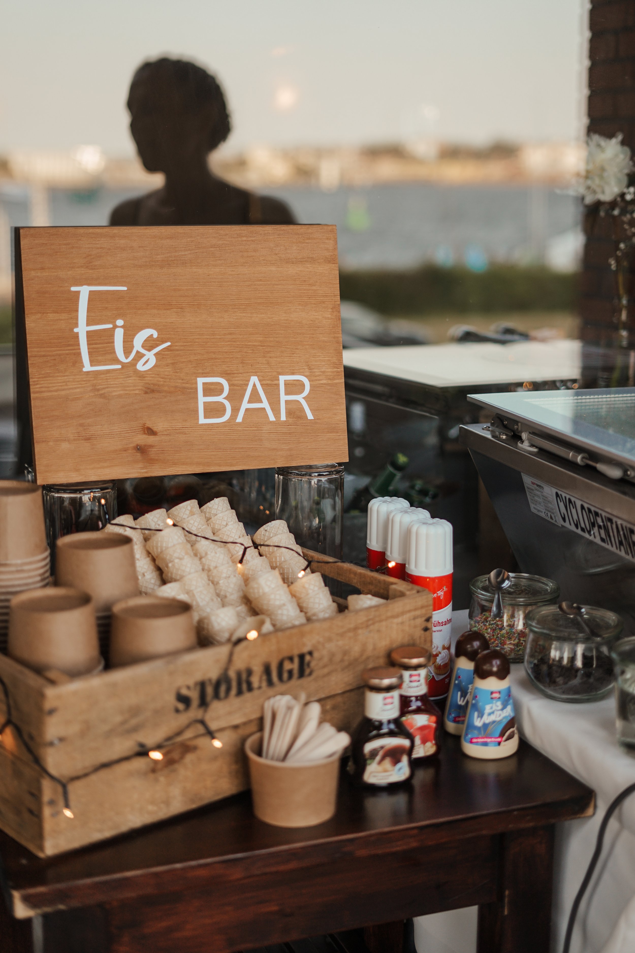 Eisbar mit Schildern und Eissorten in einer Holzkiste, auf einem Tisch mit weiteren Süßigkeiten und Dekorationen, im Hintergrund eine Scheibe mit einem menschlichen Schatten.