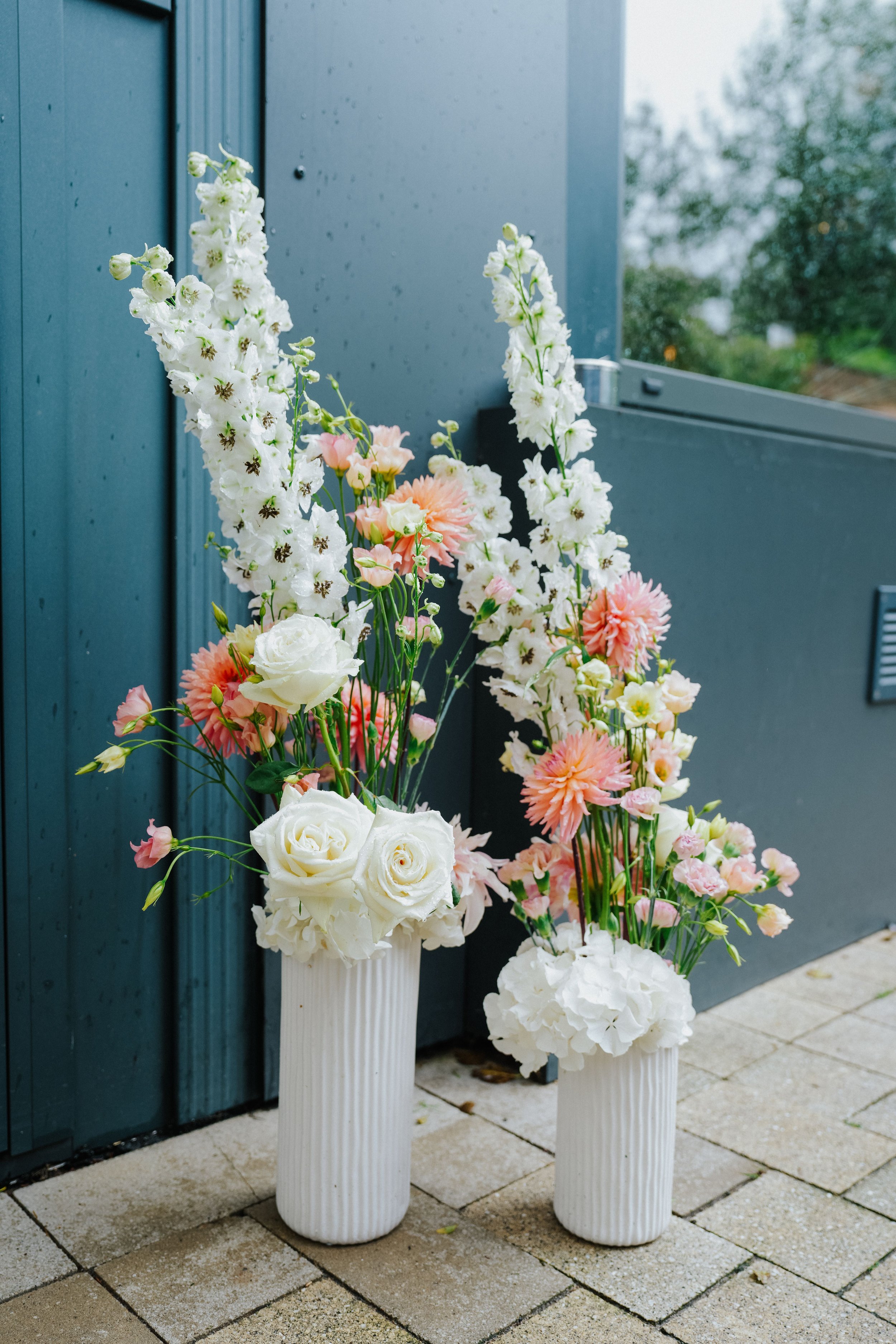 Zwei weiße Blumenkübel mit weißen und pastellfarbenen Blumen, auf einer gepflasterten Oberfläche vor einer blauen Wand.