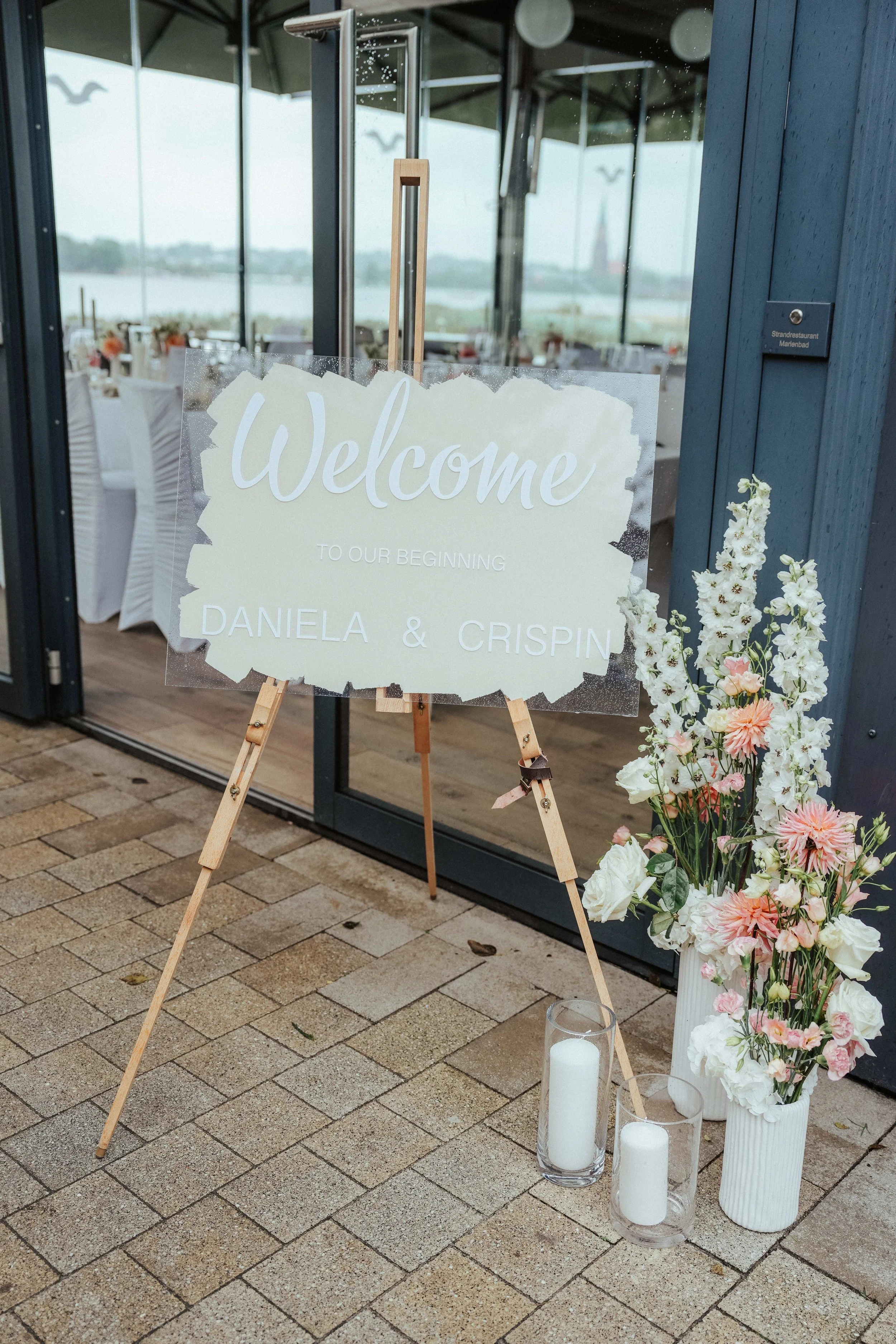 Willkommensschild für eine Hochzeitsfeier mit Blumen und Kerzen vor einem Restaurant. Sagt "Welcome" und begrüßt die Gäste für Daniela und Crispin.