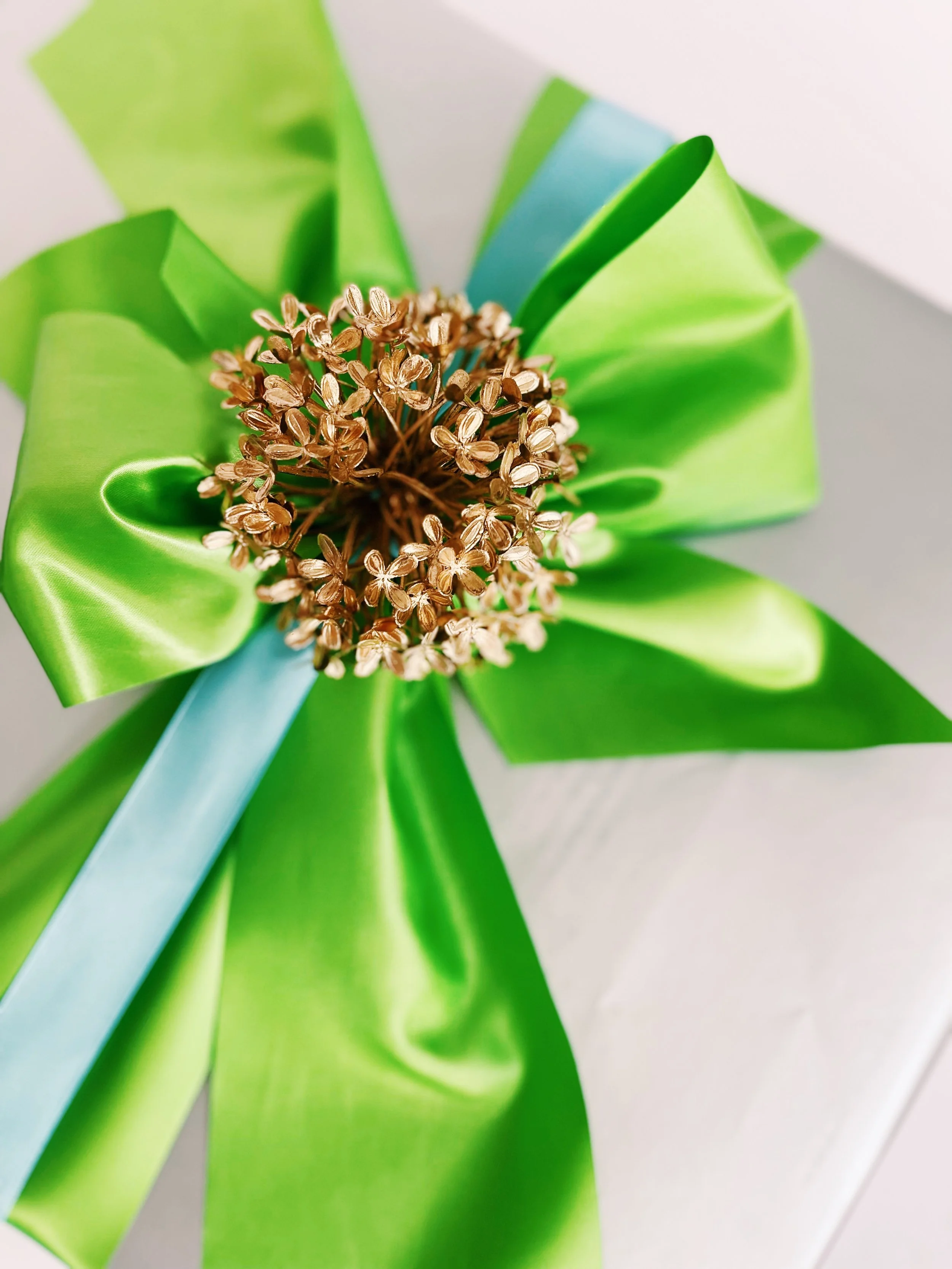 A decorative arrangement with a light blue ribbon, bright green satin ribbon, and a cluster of small dried flowers in the center, on a white background | Luxury Gift Wrapping & Custom Hampers