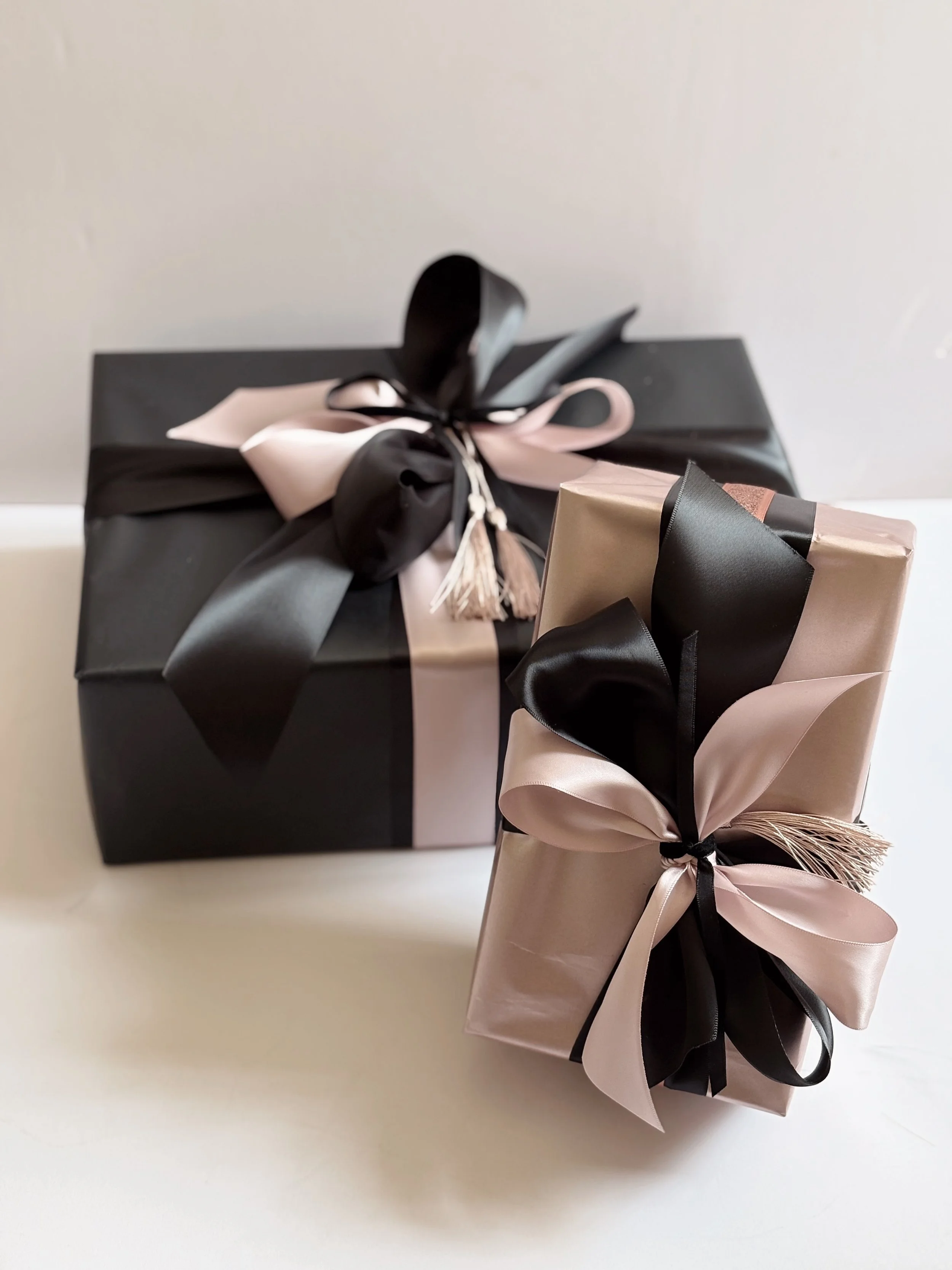 Two gift boxes wrapped with black and pink satin ribbons, decorated with bows and tassels, positioned on a white surface against a neutral background | Luxury Gift Wrapping & Custom Hampers