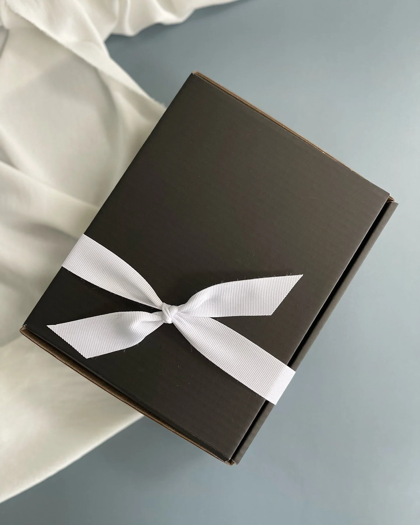 Black gift box wrapped with a white ribbon tied in a bow, placed on a light-colored cloth against a light blue background | Luxury Gift Wrapping & Custom Hampers