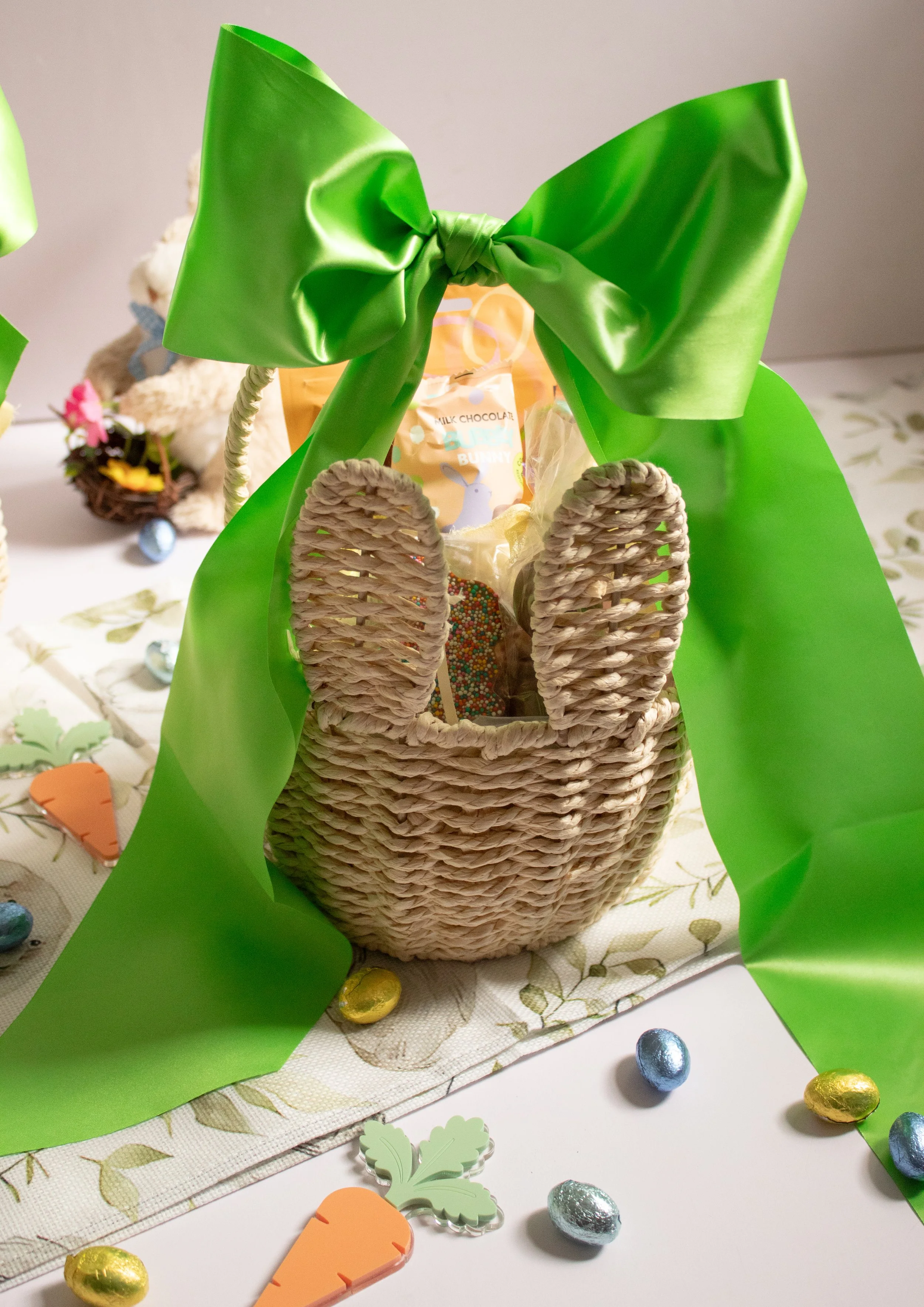 A woven basket with a large green bow on top, surrounded by small colorful foil-wrapped chocolate eggs, a paper carrot, and Easter decorations on a tablecloth | Luxury Gift Wrapping & Custom Hampers