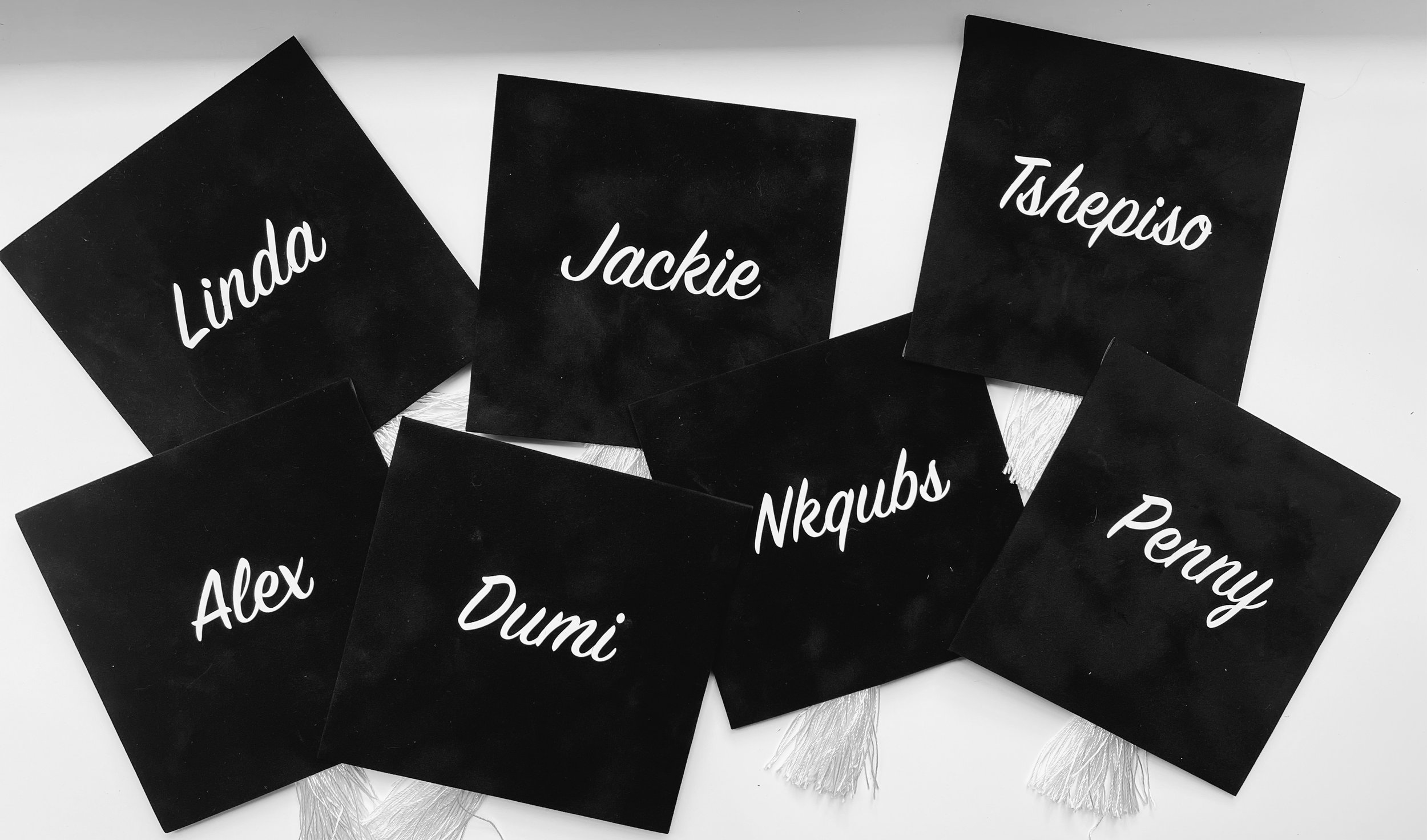 Several black paper squares with white cursive writing of various names including Linda, Jackie, Tshepiso, Alex, Dumi, Nqub, Penny, and Nkqub, scattered on a white surface | Luxury Gift Wrapping & Custom Hampers