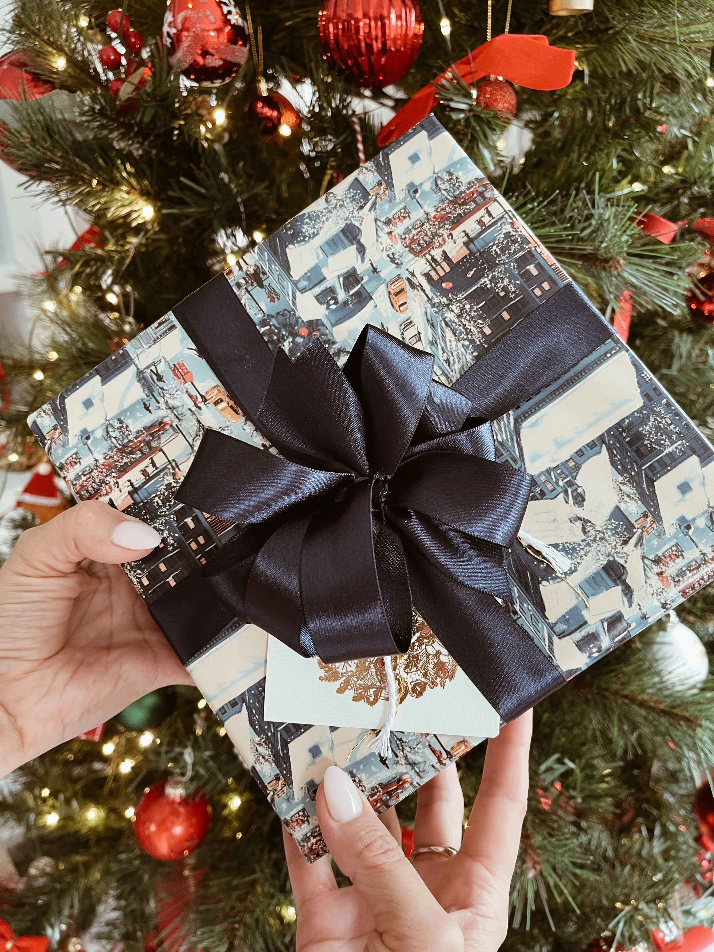 A person holding a wrapped Christmas gift with a cityscape design and a large black ribbon, in front of a decorated Christmas tree with red ornaments, lights, and ribbons | Luxury Gift Wrapping & Custom Hampers