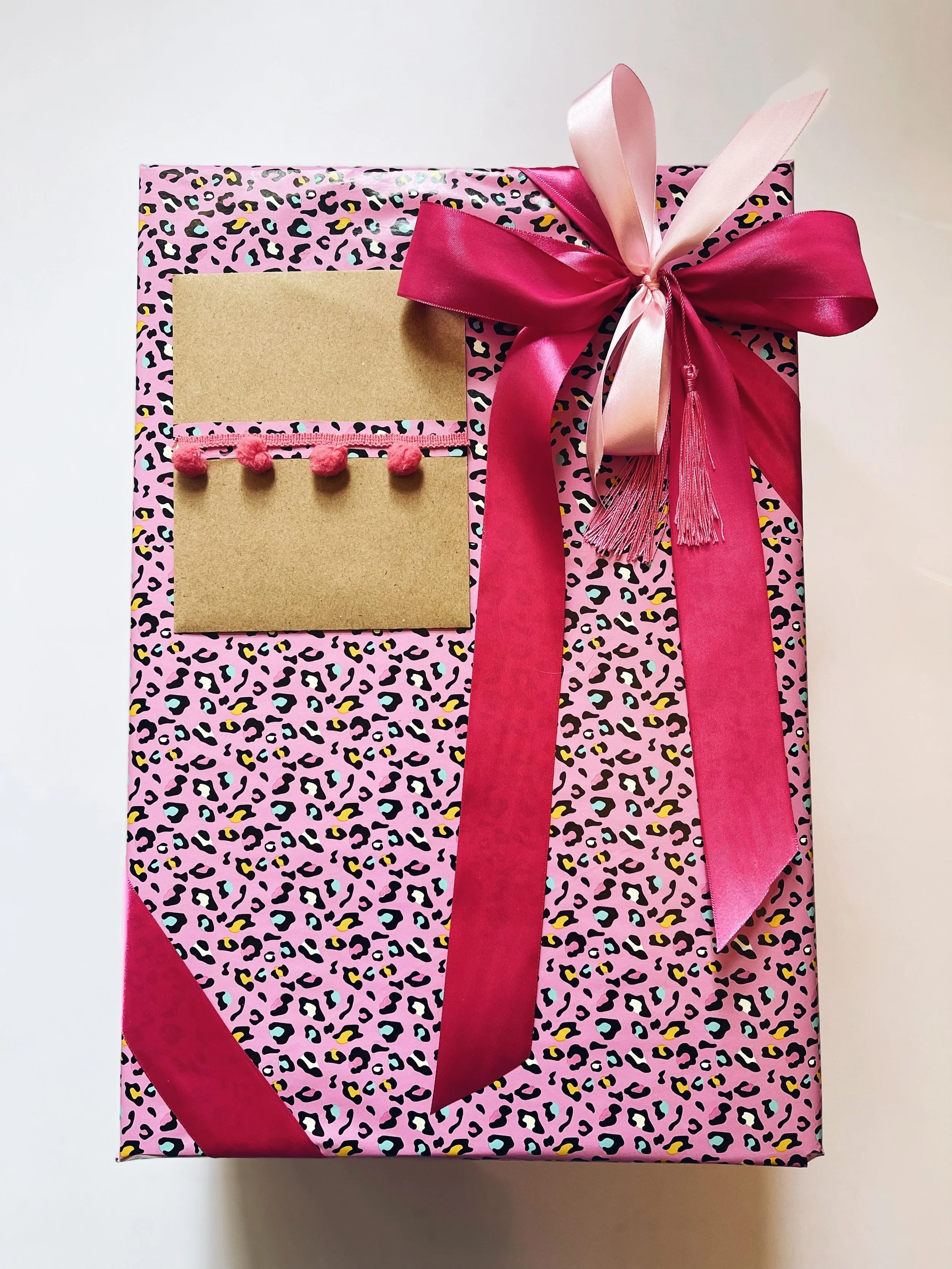 Pink leopard print gift box wrapped with a pink and dark pink satin ribbon tied in bows, with two pink note cards and pink pom-pom trim attached | Luxury Gift Wrapping & Custom Hampers.