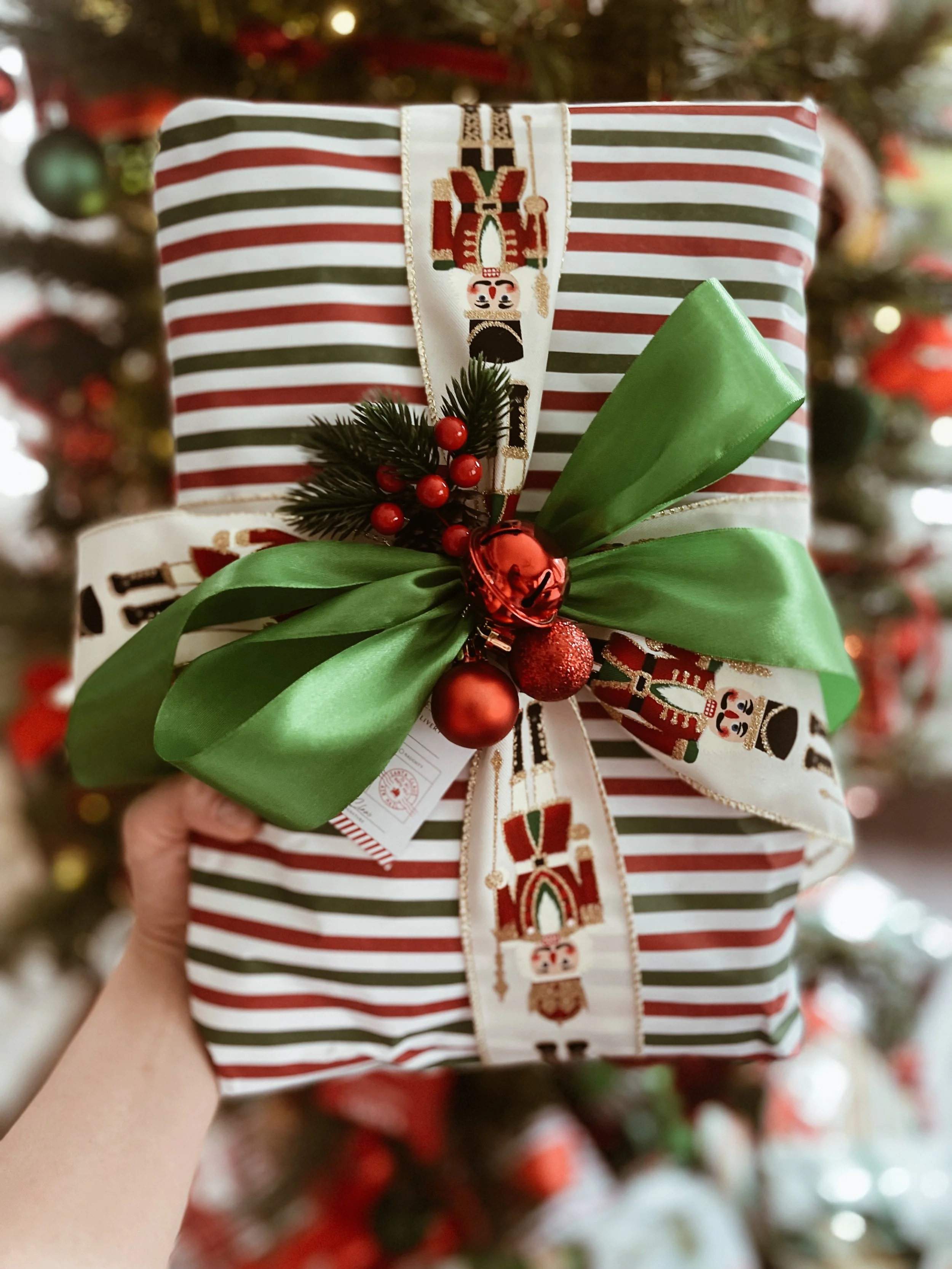 Wrapped Christmas gift with striped red, green, and white paper, topped with a large green satin bow, decorated with red ornaments, pine leaves, and berries, held in front of a Christmas tree | Luxury Gift Wrapping & Custom Hampers