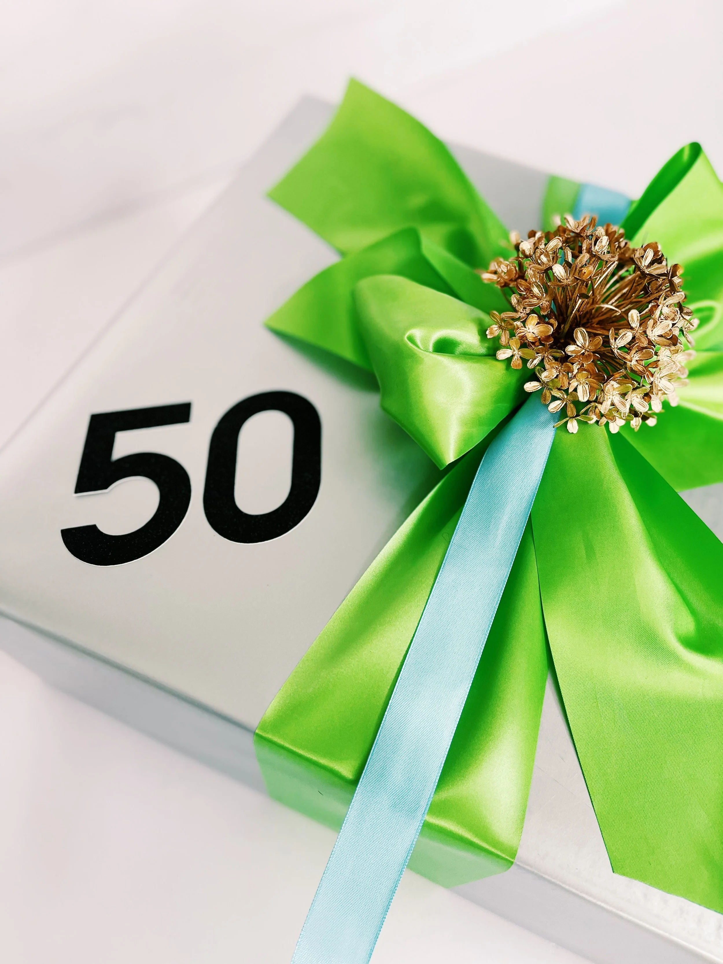 A white gift box wrapped with a green ribbon bow and a light blue ribbon, decorated with a gold-colored floral embellishment, with the number 50 printed on the top of the box | Luxury Gift Wrapping & Custom Hampers.