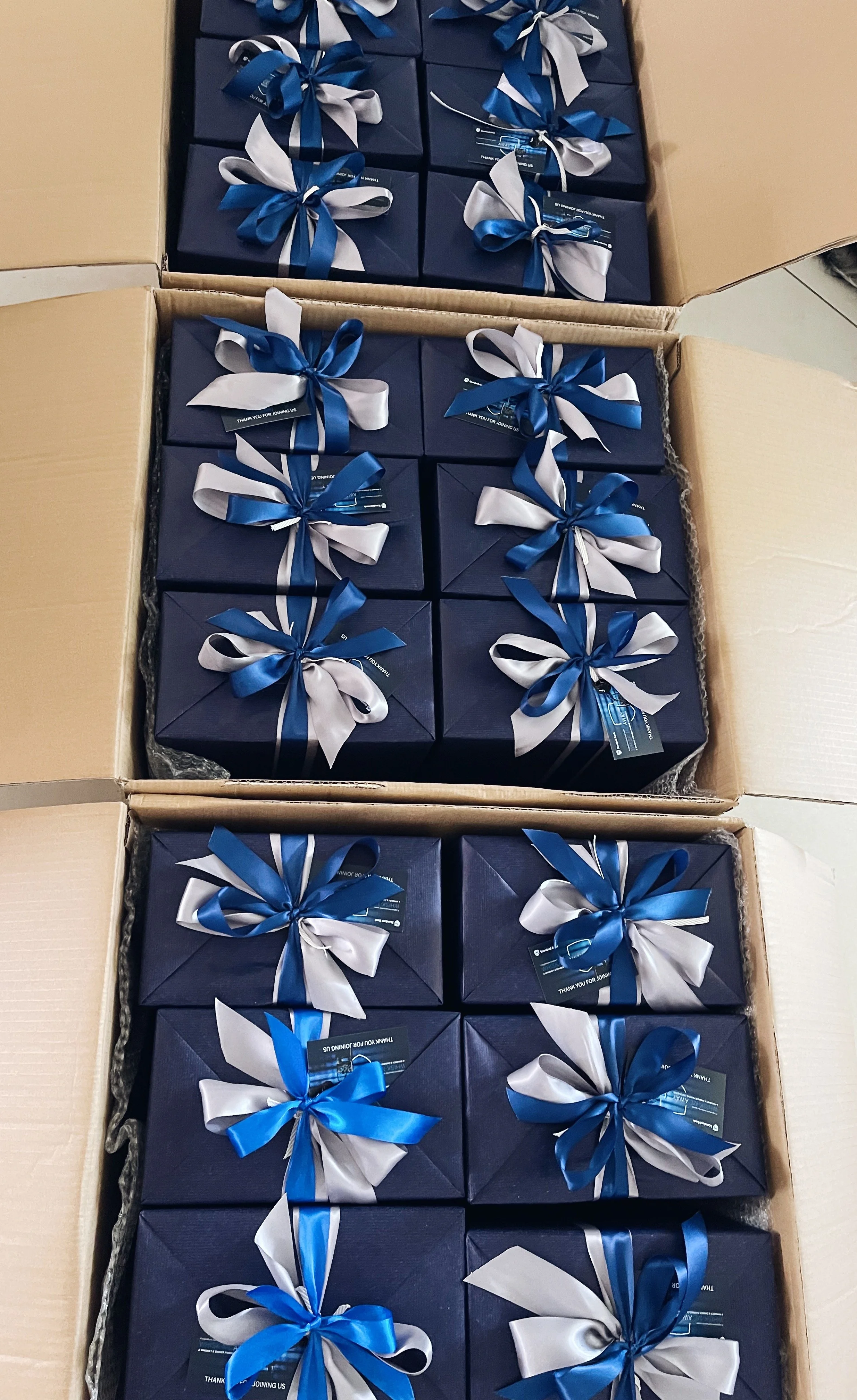 Multiple navy blue gift boxes with silver and blue ribbons stacked inside a cardboard box.
