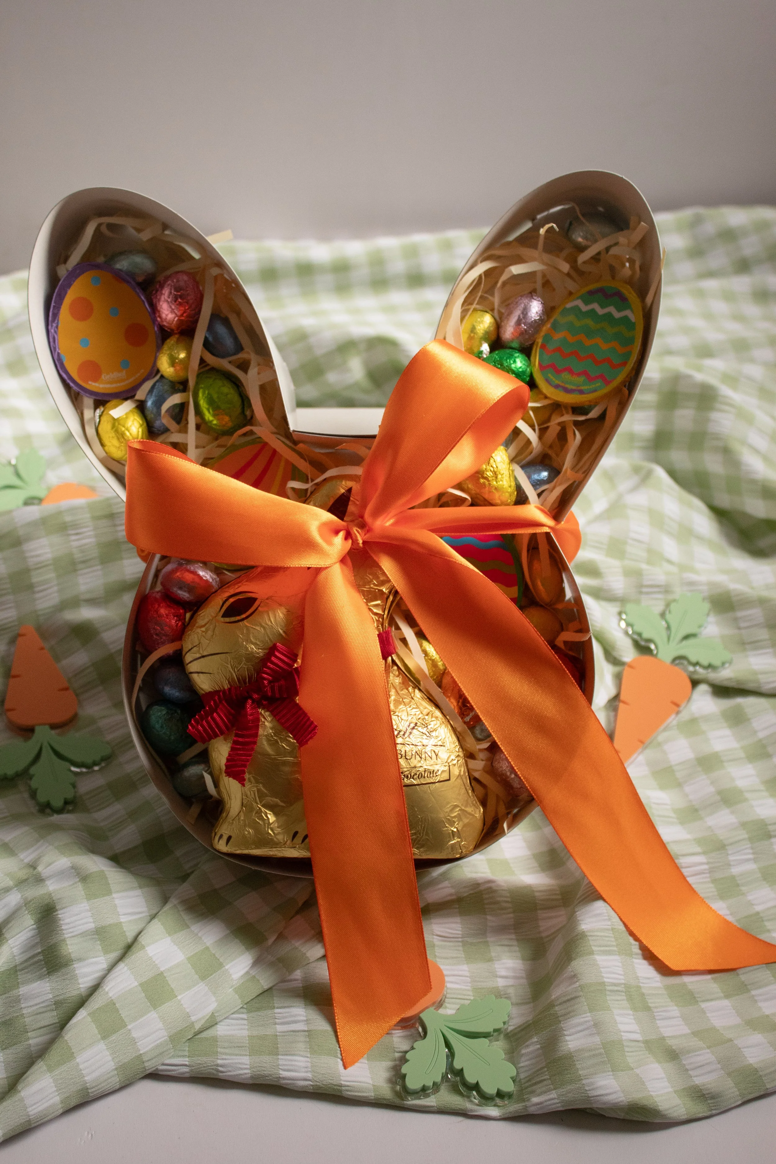 Decorative Easter basket shaped like a bunny with a large orange ribbon bow, filled with colorful Easter chocolates and candies, sitting on a green checkered cloth | Luxury Gift Wrapping & Custom Hampers