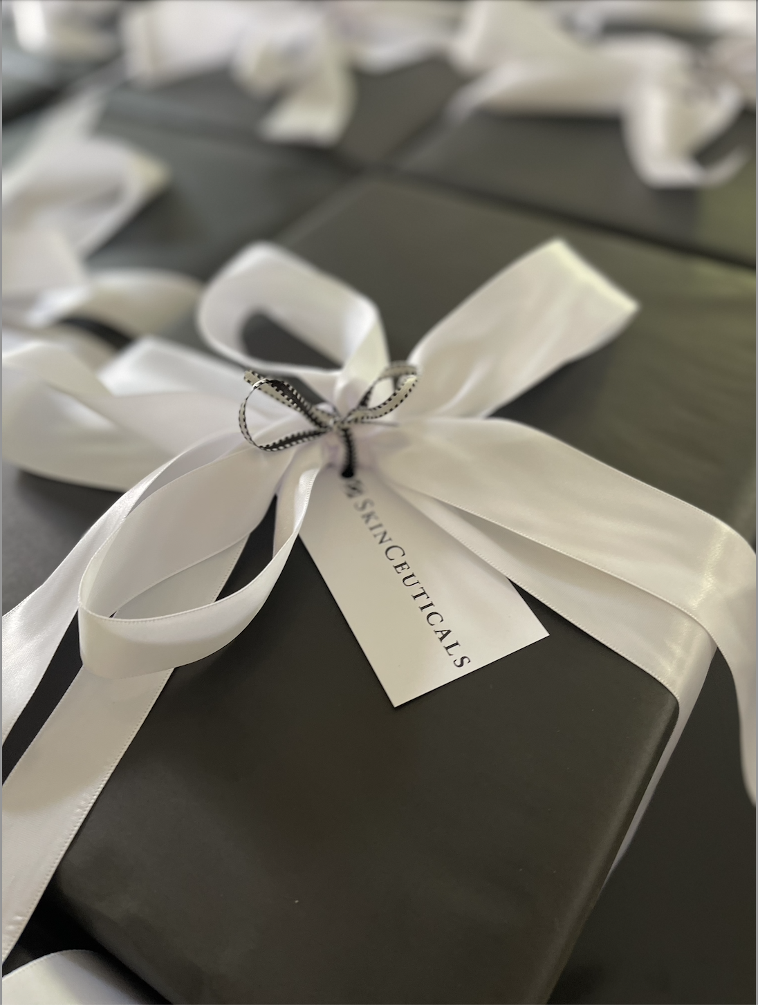 A black gift box wrapped with a white satin ribbon and bow, with a tag that reads 'SKIN CEUTICALS' and decorative black and white string | Luxury Gift Wrapping & Custom Hampers