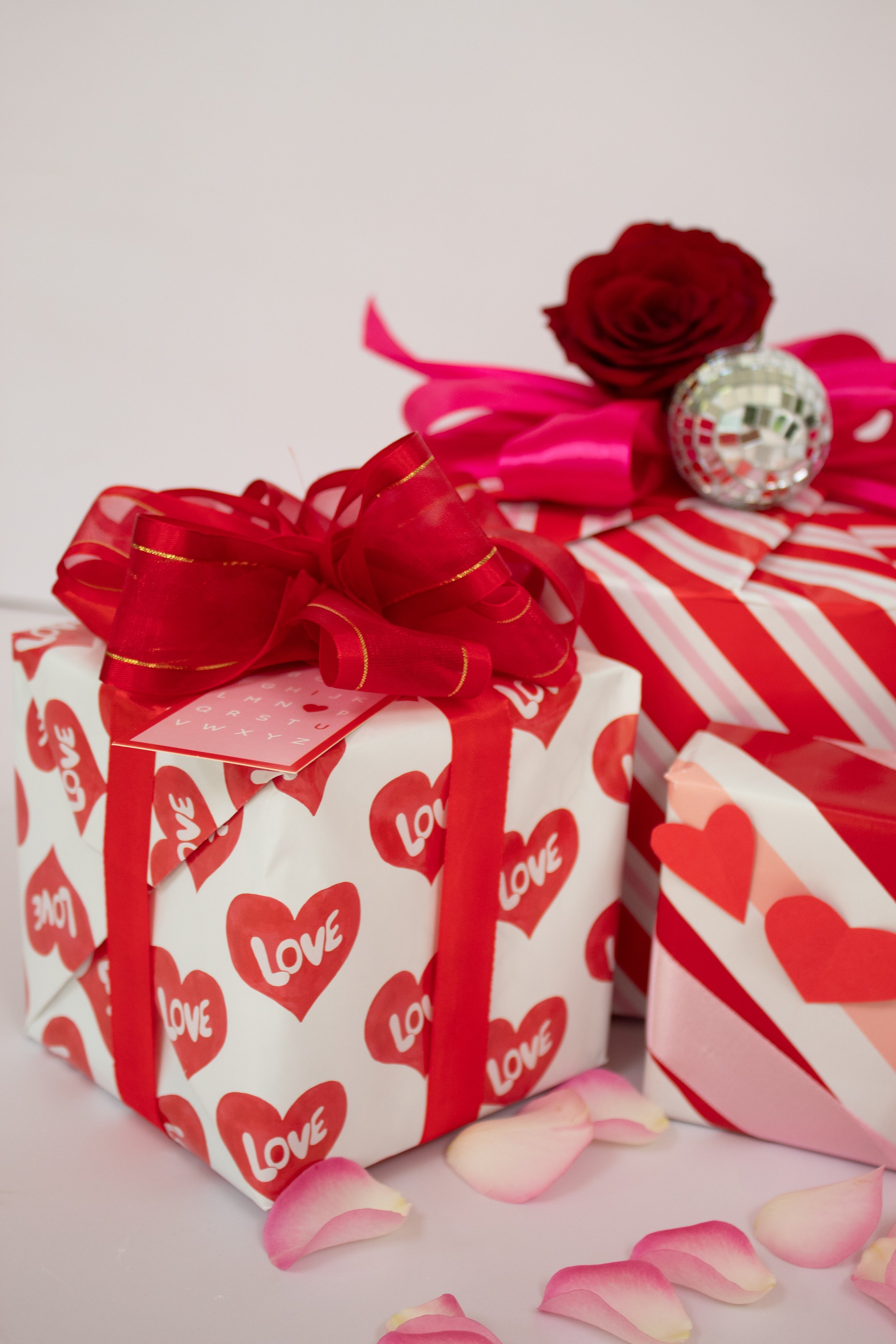 Valentine's Day gifts with red and white wrapping paper, a large red ribbon, pink rose, silver disco ball, and pink rose petals scattered on a white surface | Luxury Gift Wrapping & Custom Hampers