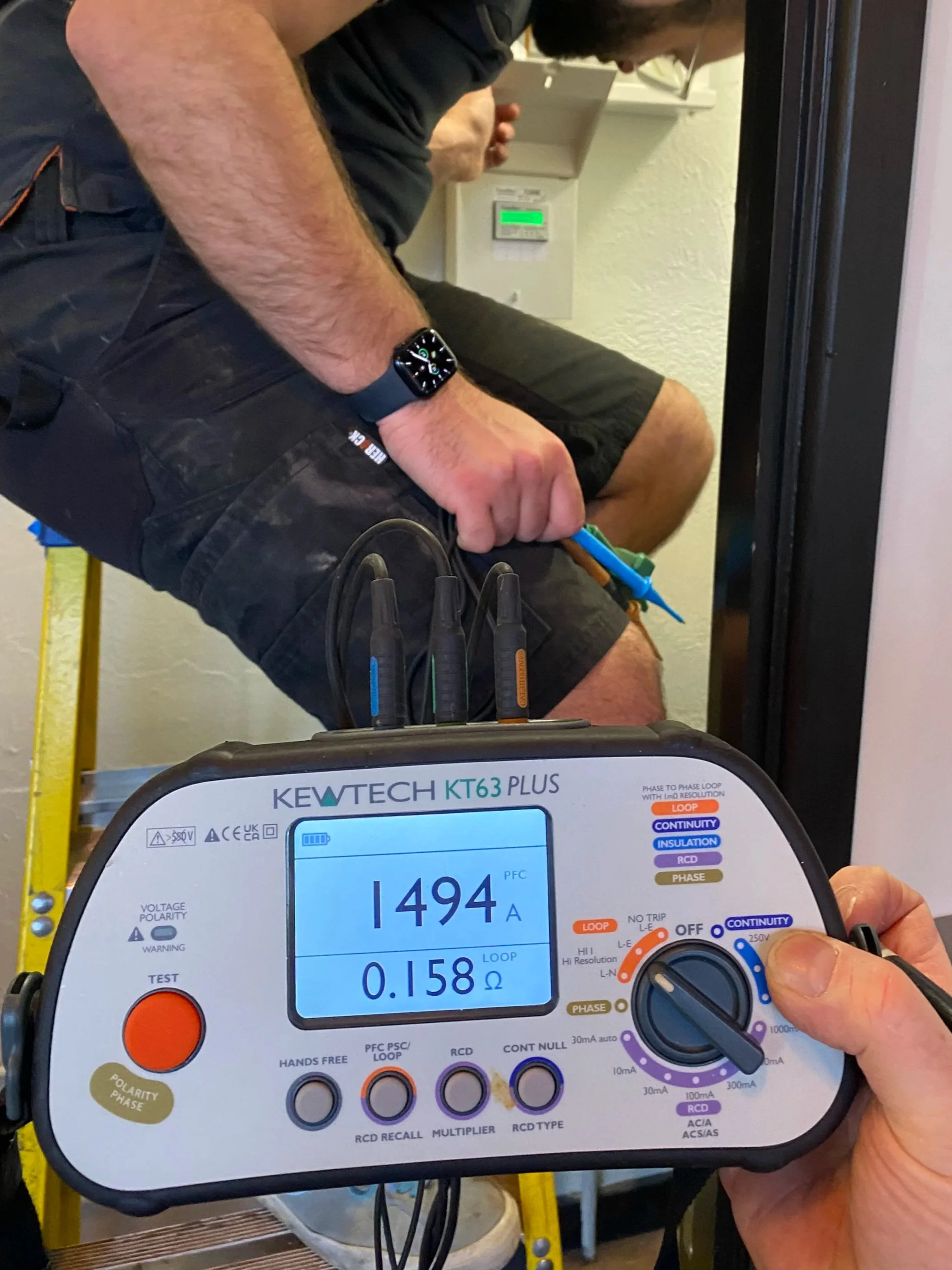 Close-up of a person using a thermal tester device, with cables connected, showing readings on the screen, while another person sits beside, holding a blue object.