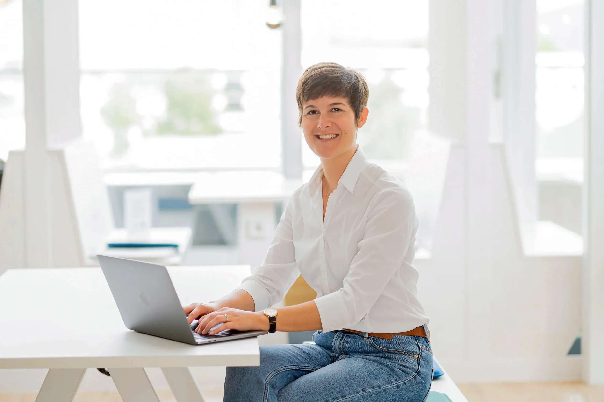 Eine Frau sitzt an einem weißen Tisch mit einem Laptop und lächelt in die Kamera, im Hintergrund helle Fenster und ein modernes Büro.