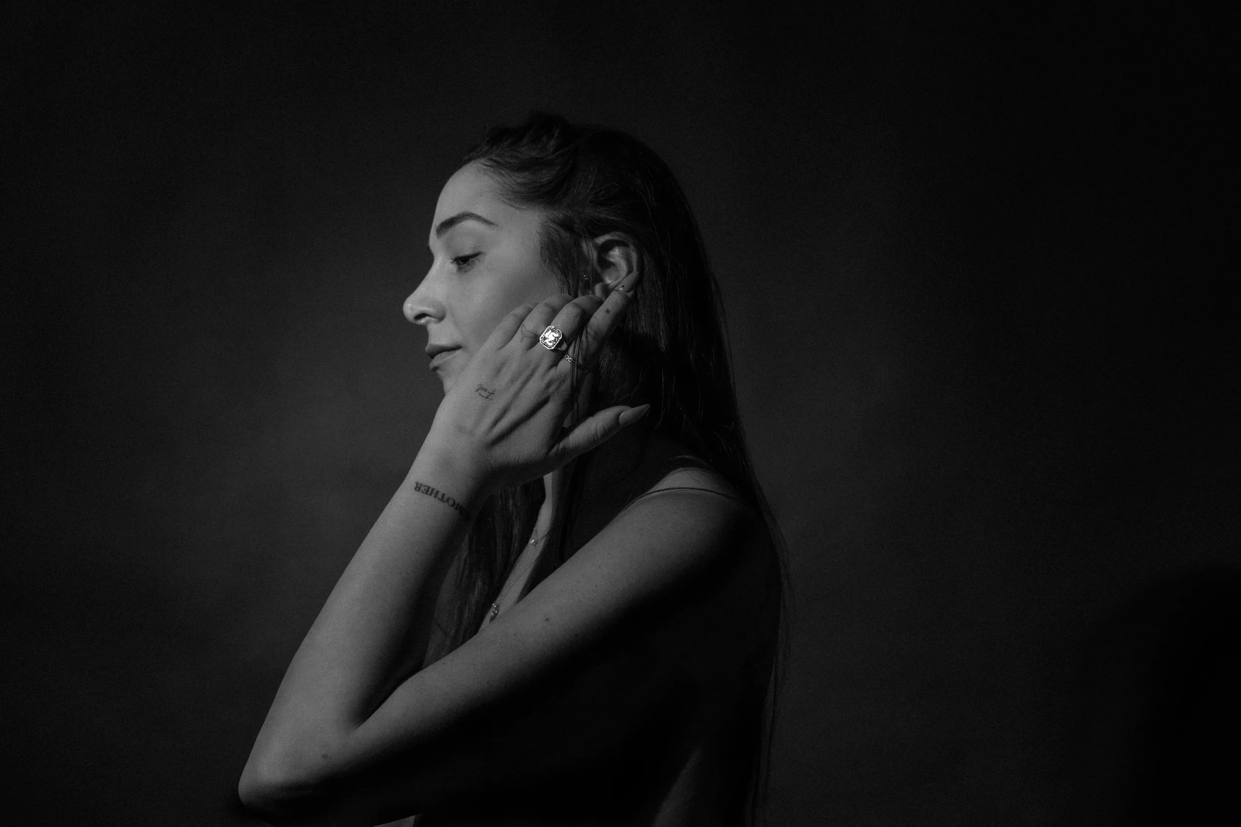 A black and white side profile photo of a woman with long hair, touching her ear gently, wearing a ring and showing tattoos on her hand and arm.