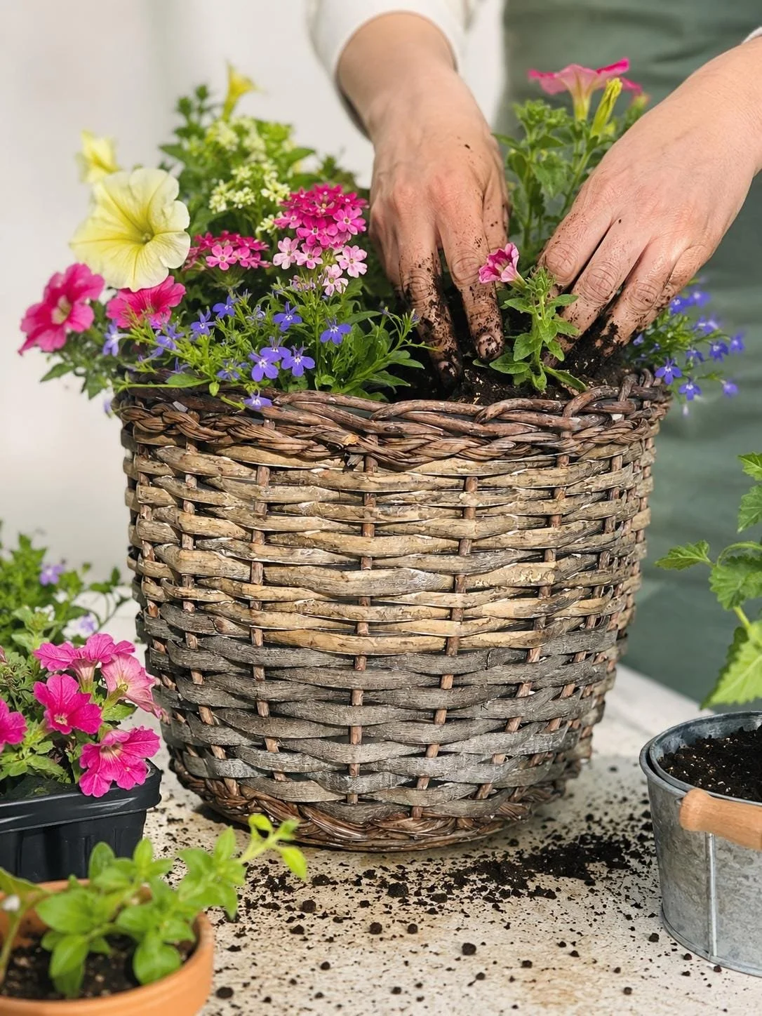 Hands planting colorful flowers in a woven basket on a gardening table.