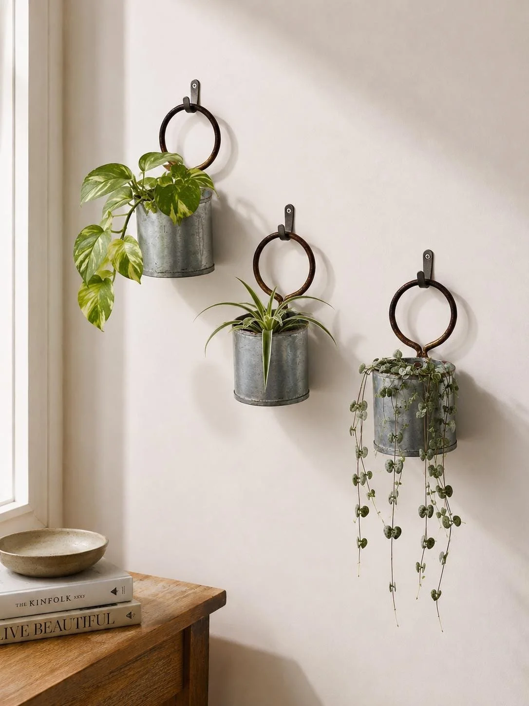 Three hanging planters with green plants on a white wall, above a wooden surface with books and a bowl.