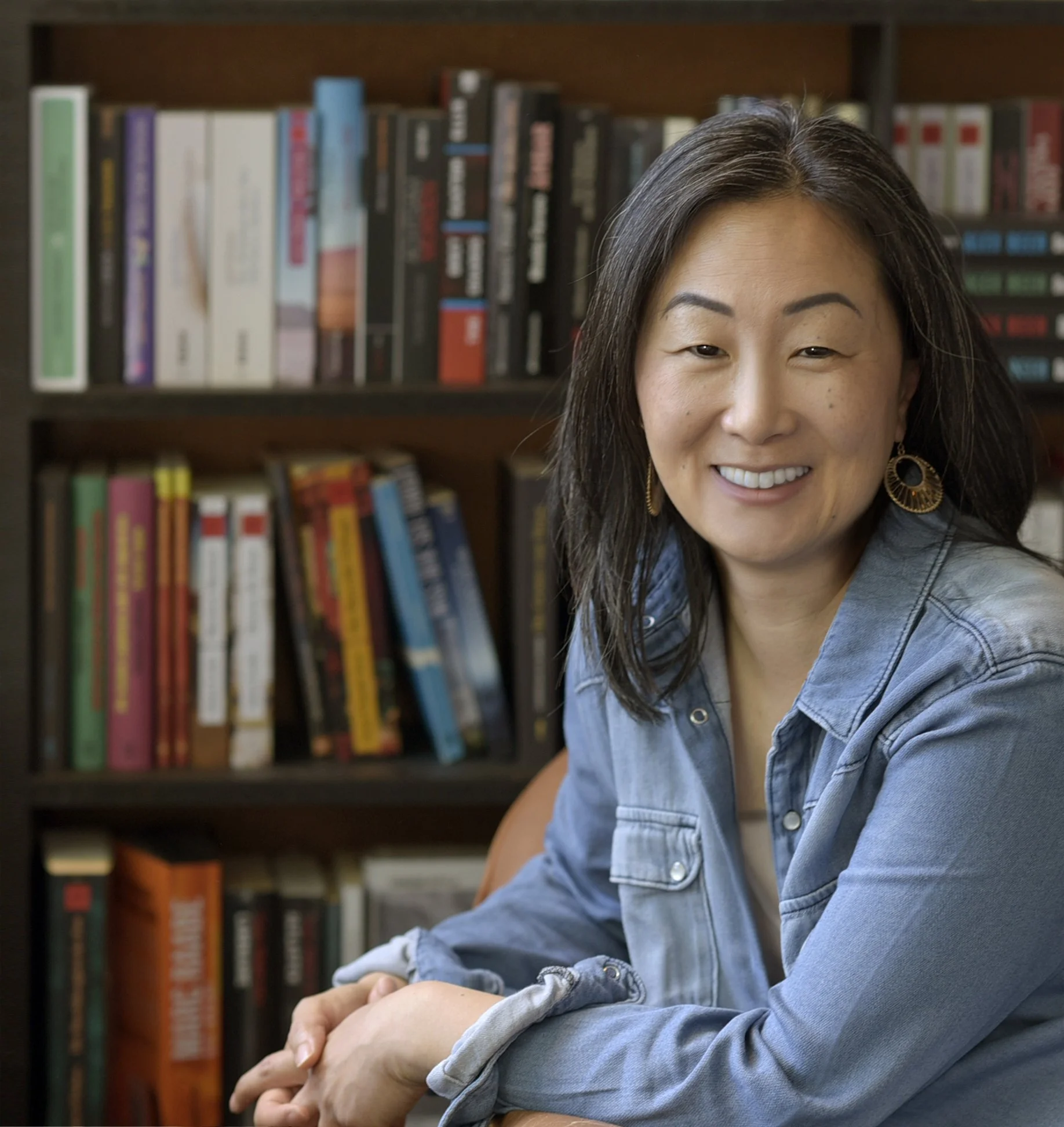 Frau mit dunklen Haaren, die ein blaues Jeanshemd trägt, sitzt vor einem Bücherregal und lächelt.