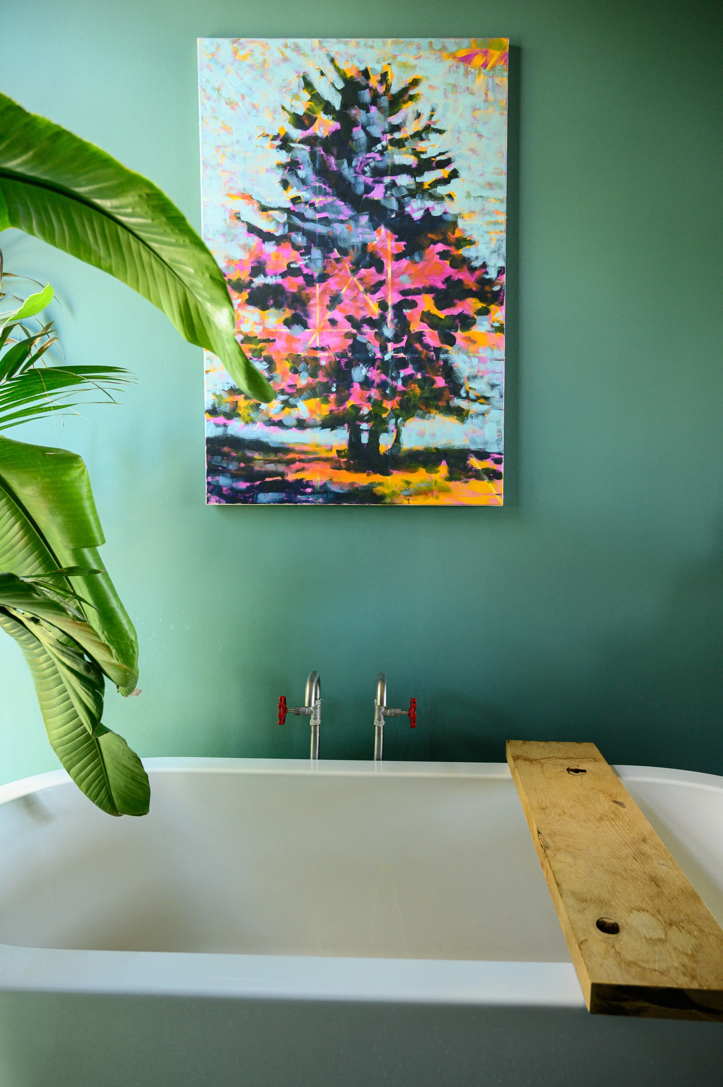 Ein Badebereich mit grüner Wand, einem weißen Badewannen-Style-Bad, zwei Wasserhähnen mit roten Griffen, einem Holzbrett auf der Wanne, einem großen, abstrakten Baumkunstwerk an der Wand und einer großen grünen Zimmerpflanze