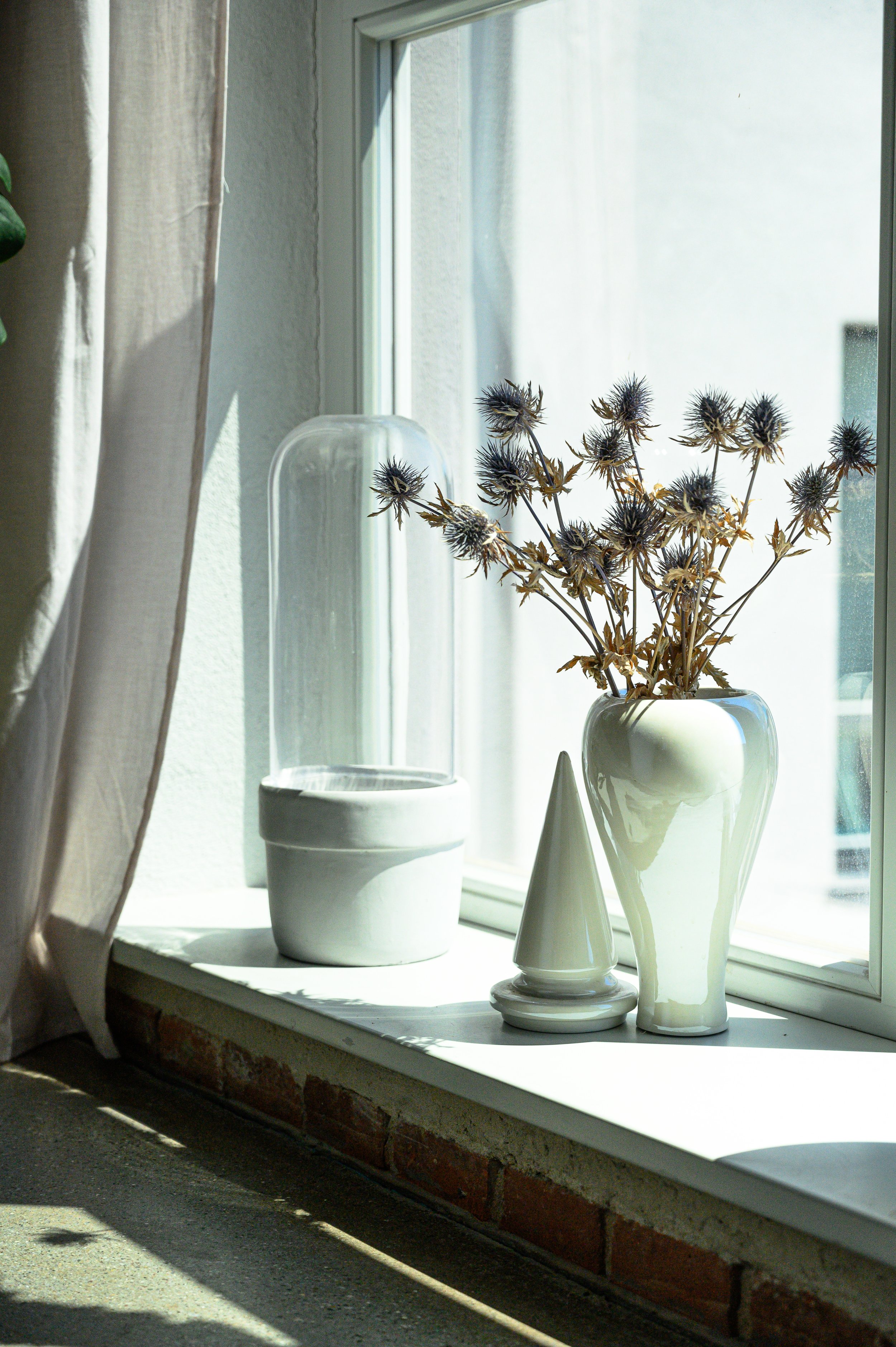 Weiße Vase mit vertrockneten Disteln auf Fenstersims, daneben ein Glasfackel und eine weiße Skulptur in Sonnenlicht.