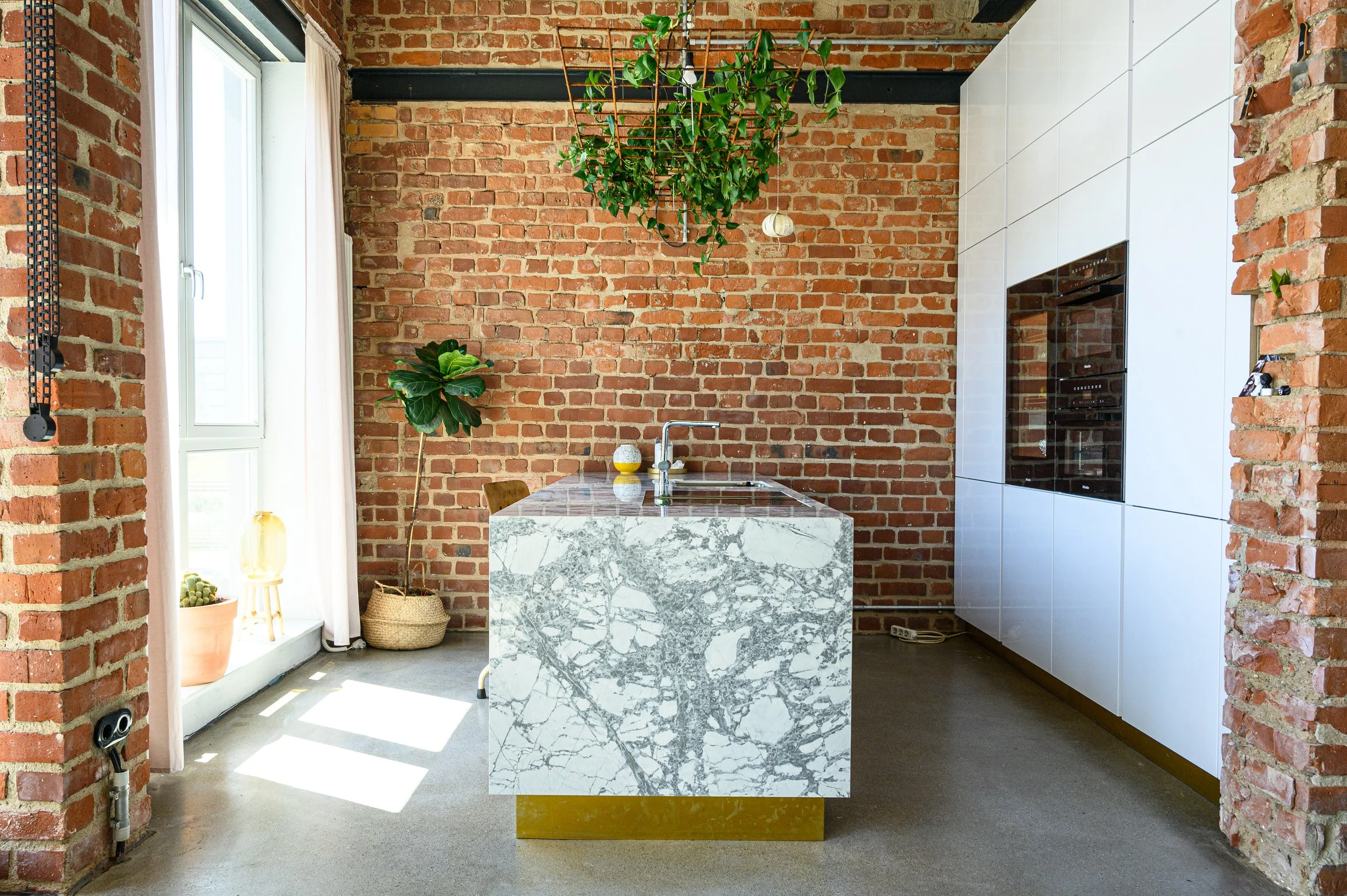 Moderne Küche mit Backsteinwänden, einem weißen Küchenschrank, einem Marmor-Insel mit Wasserhahn und Pflanzen neben einem großen Fenster.