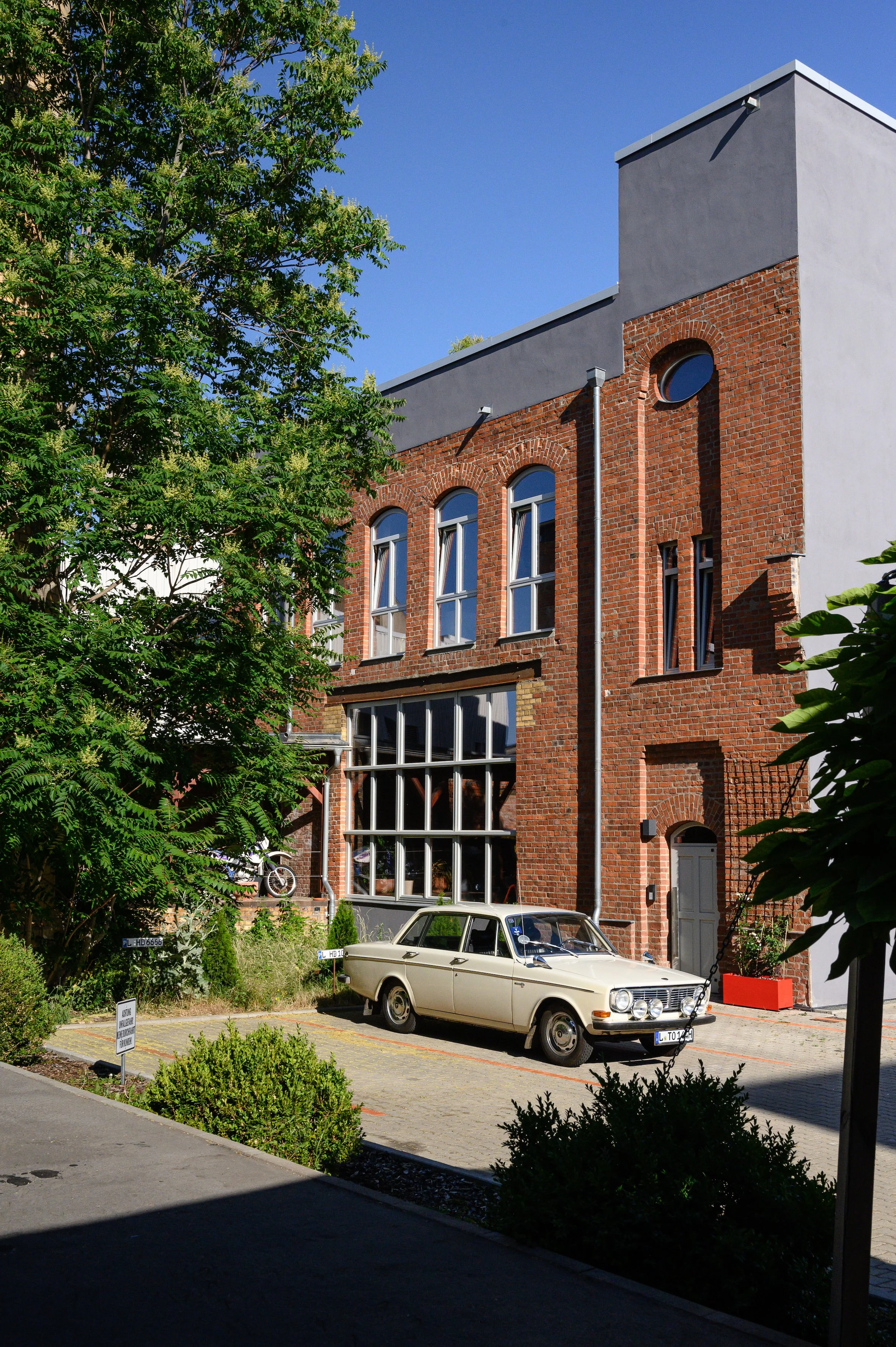 Ein alter weißer Autoklassiker, geparkt vor einem roten Backsteingebäude mit großen Fenstern, umgeben von grünen Büschen und einem Baum, bei sonnigem Himmel.